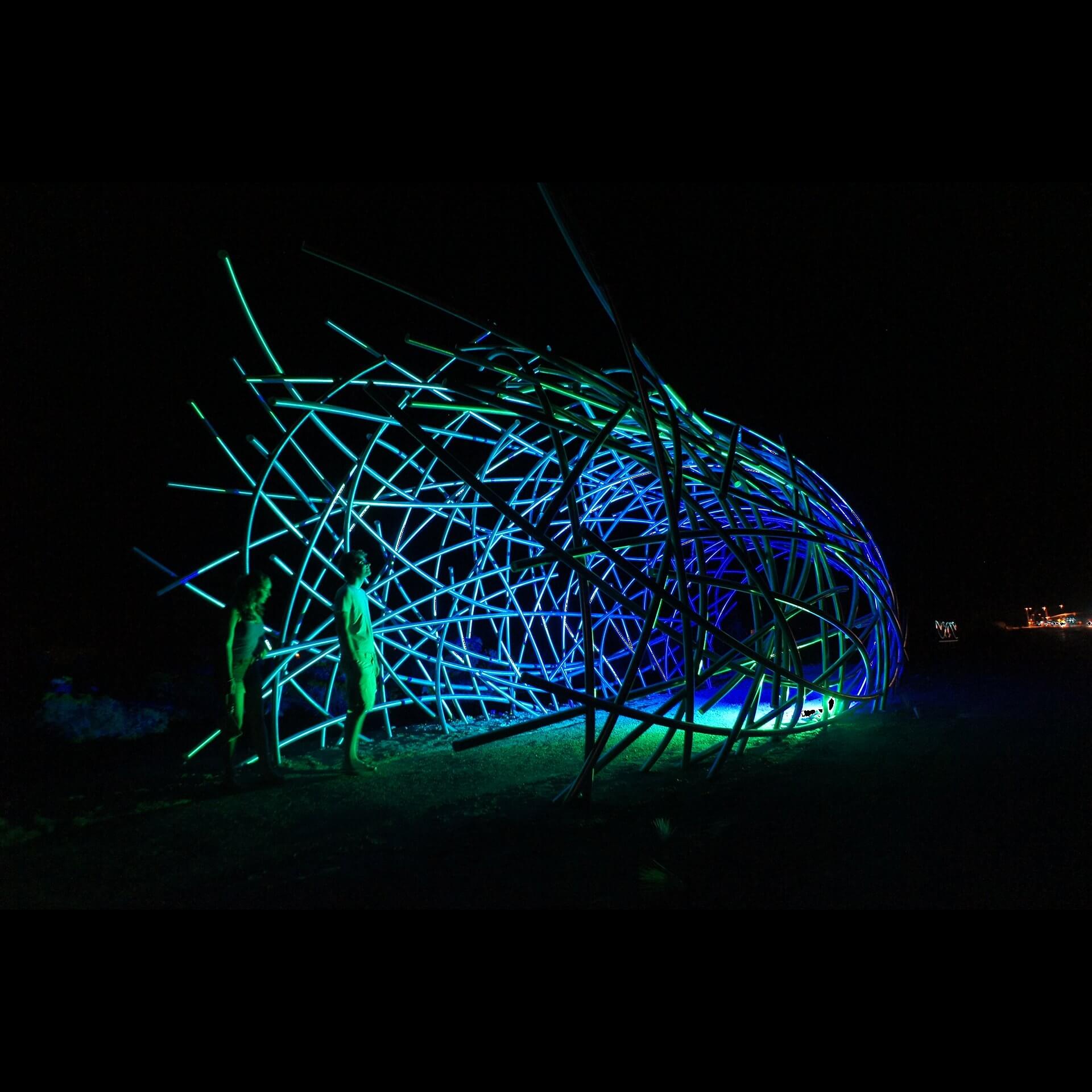 Night scene featuring two people standing near an illuminated art installation made of intertwining metal rods. The structure, a creation by renowned sculpture artist Blessing Hancock, is lit with green and blue lighting, creating a vibrant glow against the dark background.