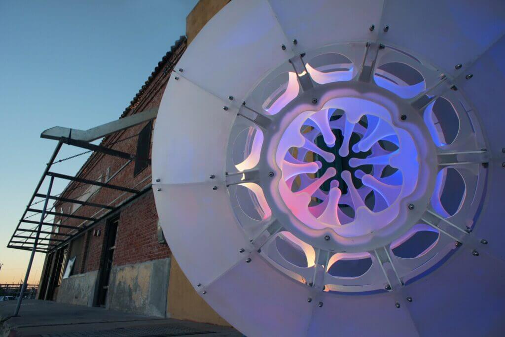 A large, futuristic circular structure with intricate, luminous patterns stands in front of an industrial-style building made of brick and metal. Designed by renowned sculpture artist Blessing Hancock, the structure is lit with blue, purple, and pink lights under a clear sky.