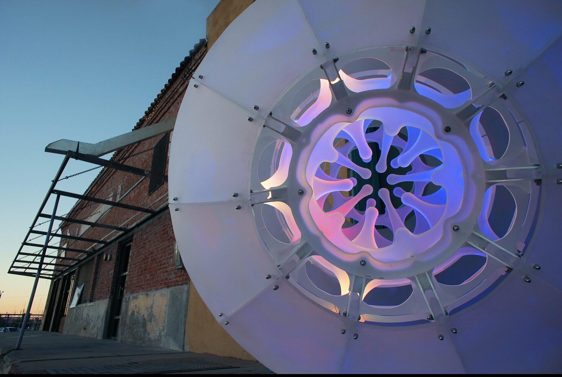 A close-up photo of a large, circular, translucent art installation by Blessing Hancock, featuring intricate patterns illuminated with blue and purple lights. The background shows a brick building with a metal awning and a clear evening sky.