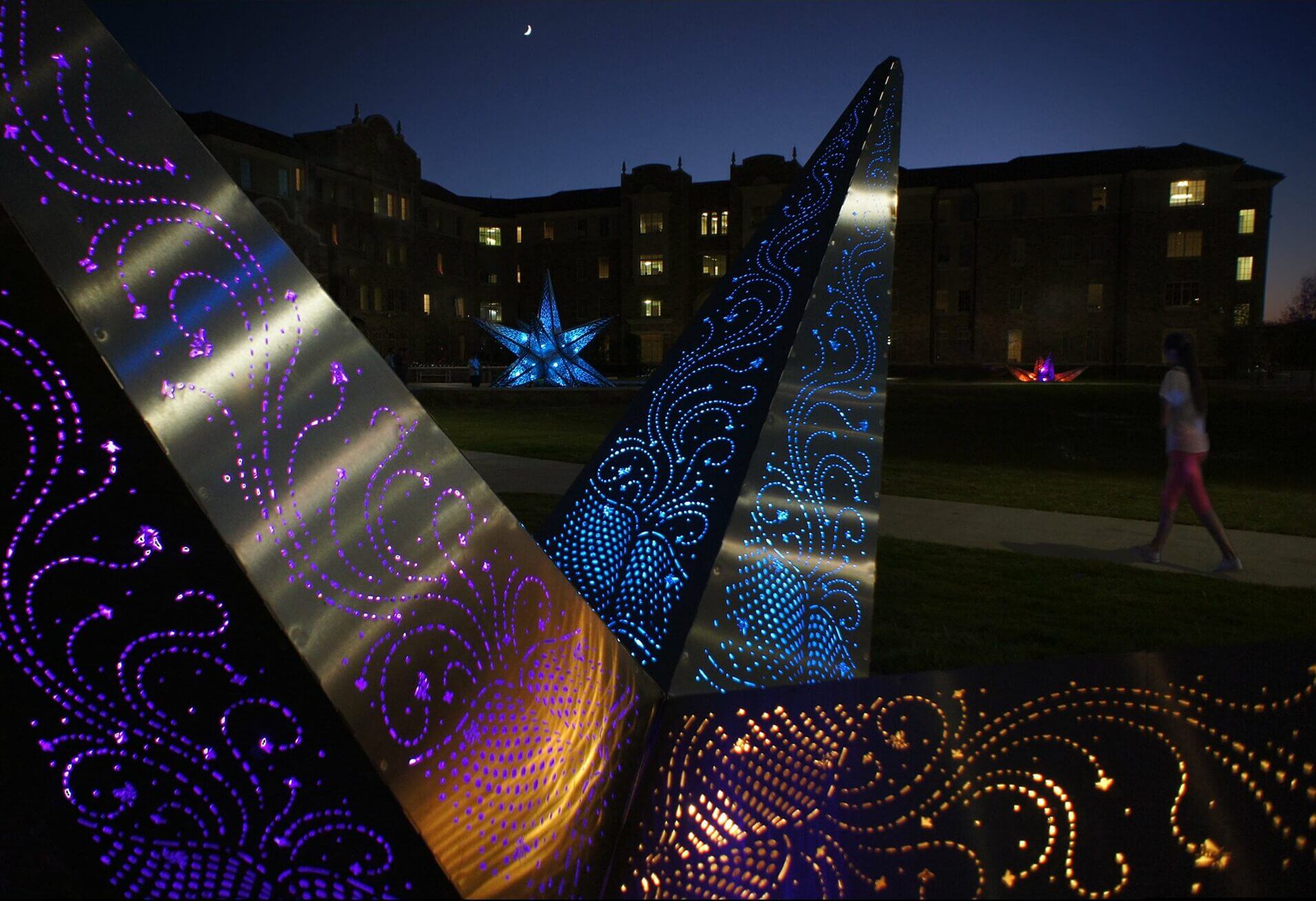 A nighttime scene shows the illuminated sculptures of Blessing Hancock, renowned sculpture artist, with intricate patterns glowing in purple and blue. A crescent moon is visible in the sky. A person walks along a path beside the sculptures, while a building with lighted windows stands in the background.