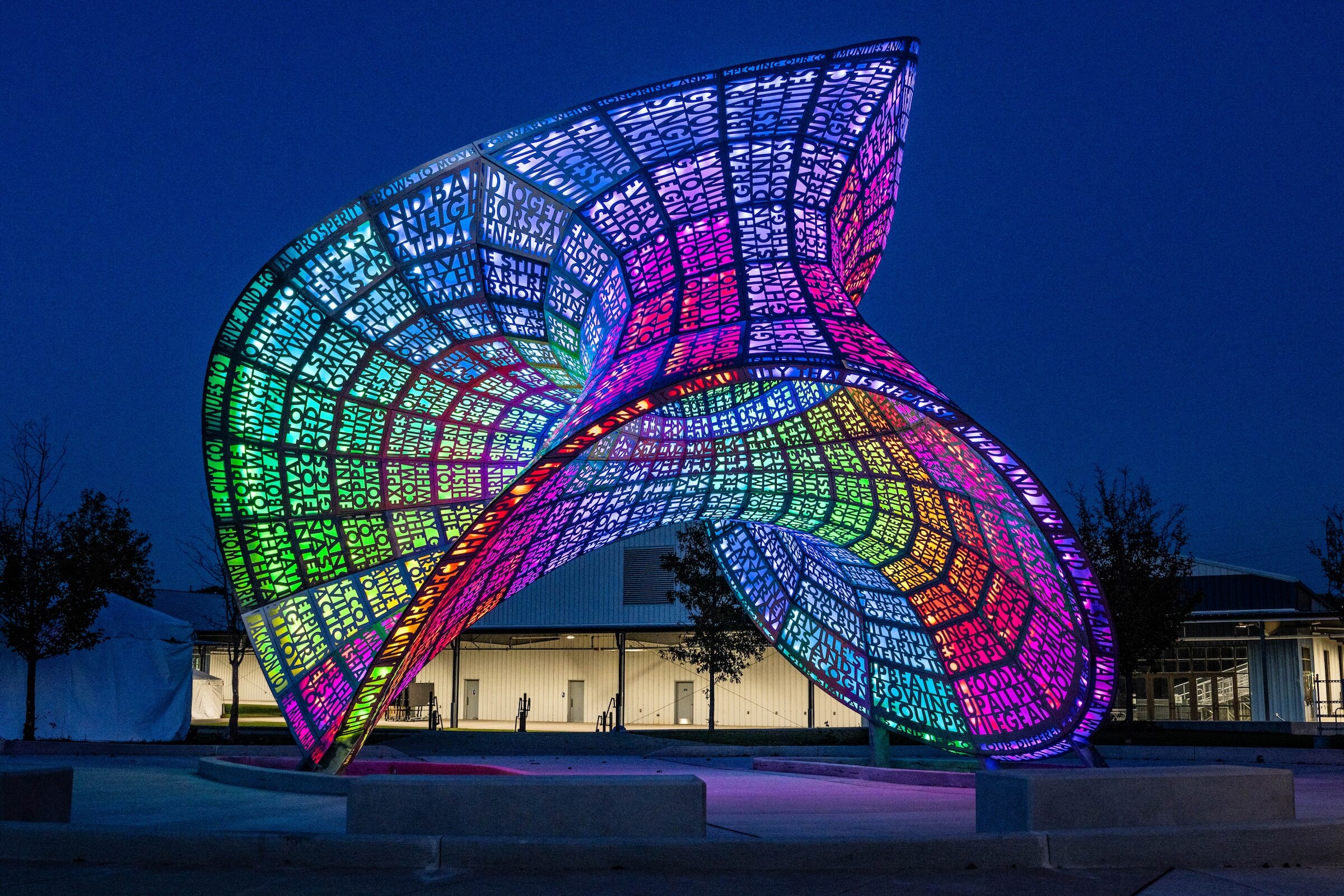 A vibrant, large-scale Loqui art sculpture illuminated at night with multicolored, geometric patterns. The structure is an intricate, twisting form with a mesh-like surface, glowing brightly against a dark blue sky. A building and trees are visible in the background.