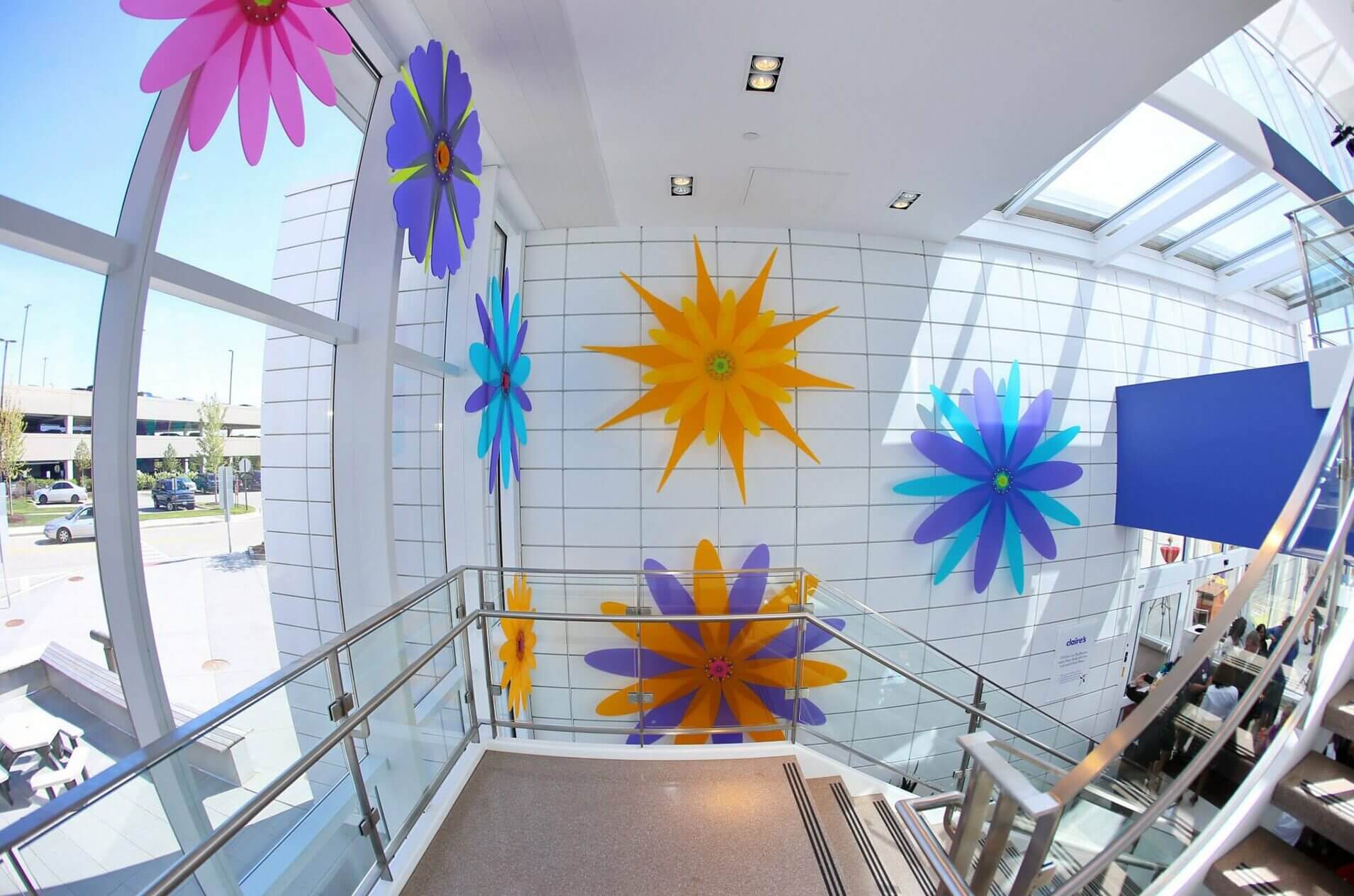 A bright, modern indoor space with a wall decorated by large, vibrant flower sculptures in purple, blue, yellow, and pink. Sunlight streams through skylights while Blessing Hancock's artistry shines through. Outside, a parking area is partially visible through tall windows.