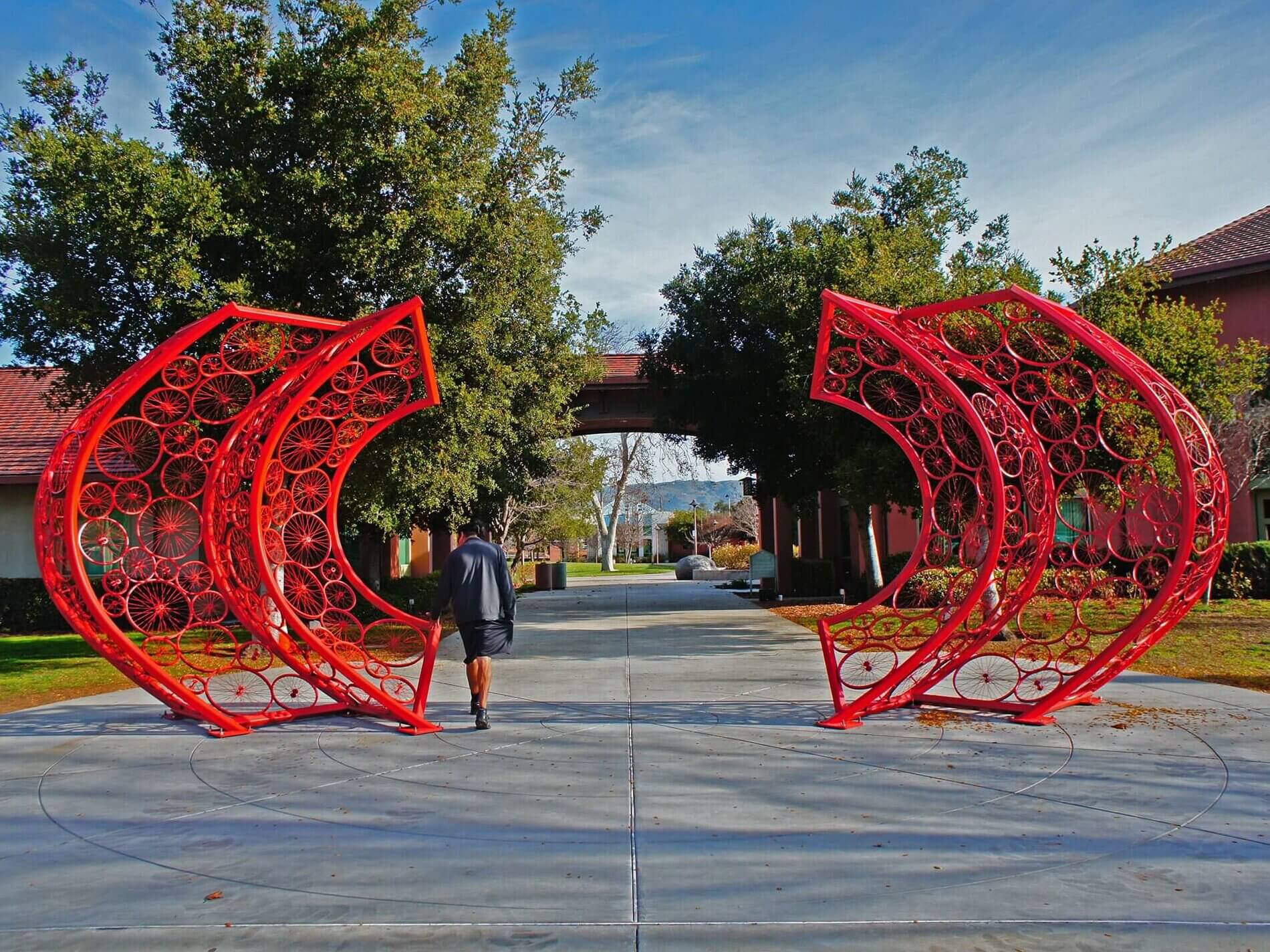 A person wearing a black jacket walks through a unique red archway sculpture made of circular patterns by Blessing Hancock, located on a paved path lined with green trees and red-roofed buildings. The sky is clear with scattered clouds.