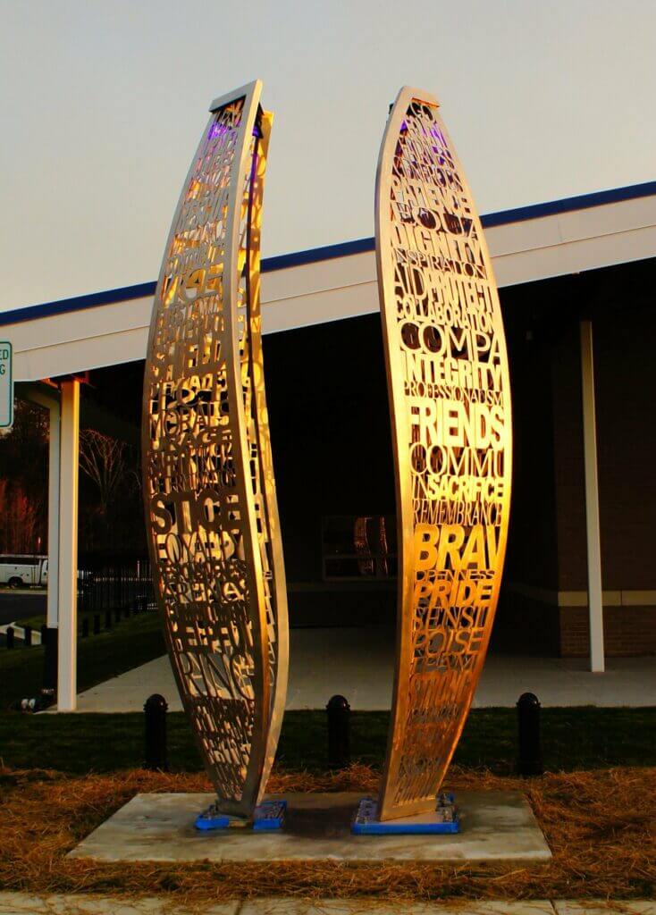 A modern outdoor sculpture by Blessing Hancock, consisting of two vertical, curving metallic panels standing parallel to each other. Each panel is intricately cut to form words such as "INTEGRITY," "FRIENDS," and "BRAVE." The sculpture stands in front of a building with a covered entrance.