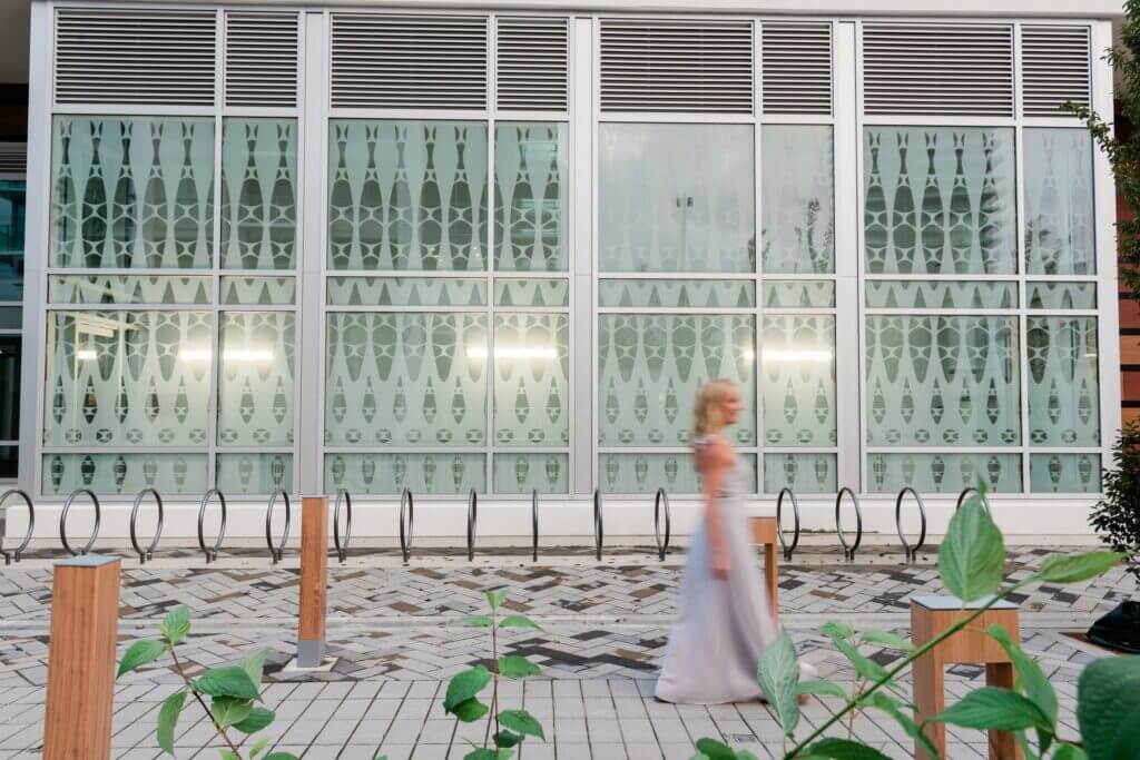 A woman in a flowing white dress walks past a modern building with large patterned glass windows and a row of bike racks. The ground is made up of a mosaic of rectangular tiles, and there are some green plants in the foreground, echoing the artistry reminiscent of Blessing Hancock's sculptures.