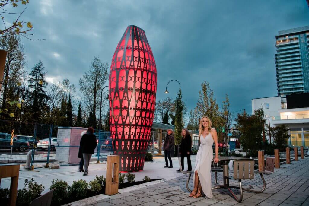 A woman in a white dress stands near a modern art sculpture by Blessing Hancock, illuminated with red lights. The scene is set against a twilight urban landscape with a few people walking, surrounded by buildings and trees.