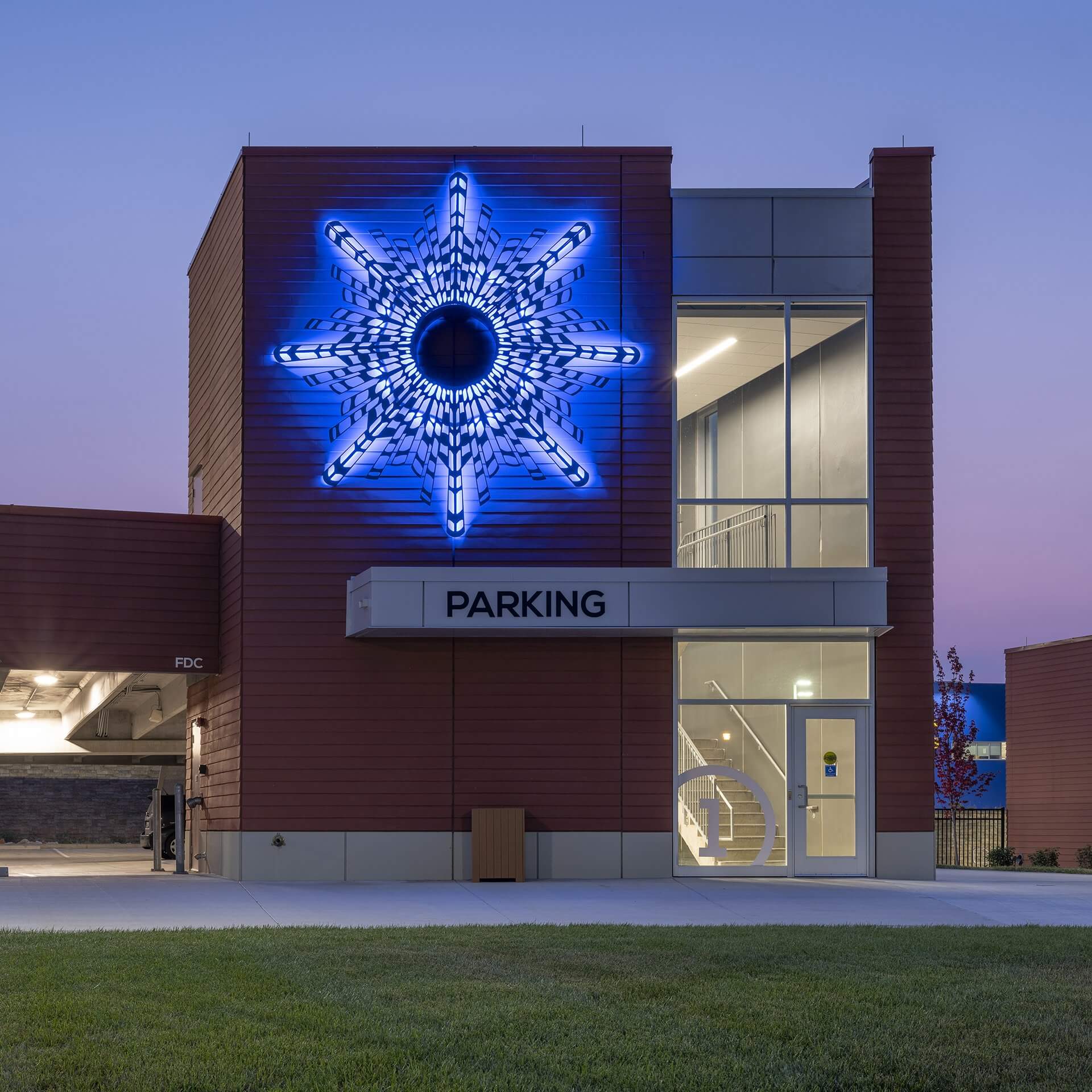 A modern parking structure with a striking blue neon star-like art installation by sculpture artist Blessing Hancock on the exterior wall. The building has a glass stairwell illuminated from within and a sign that reads "PARKING" above the entrance. The grass lawn is in the foreground.