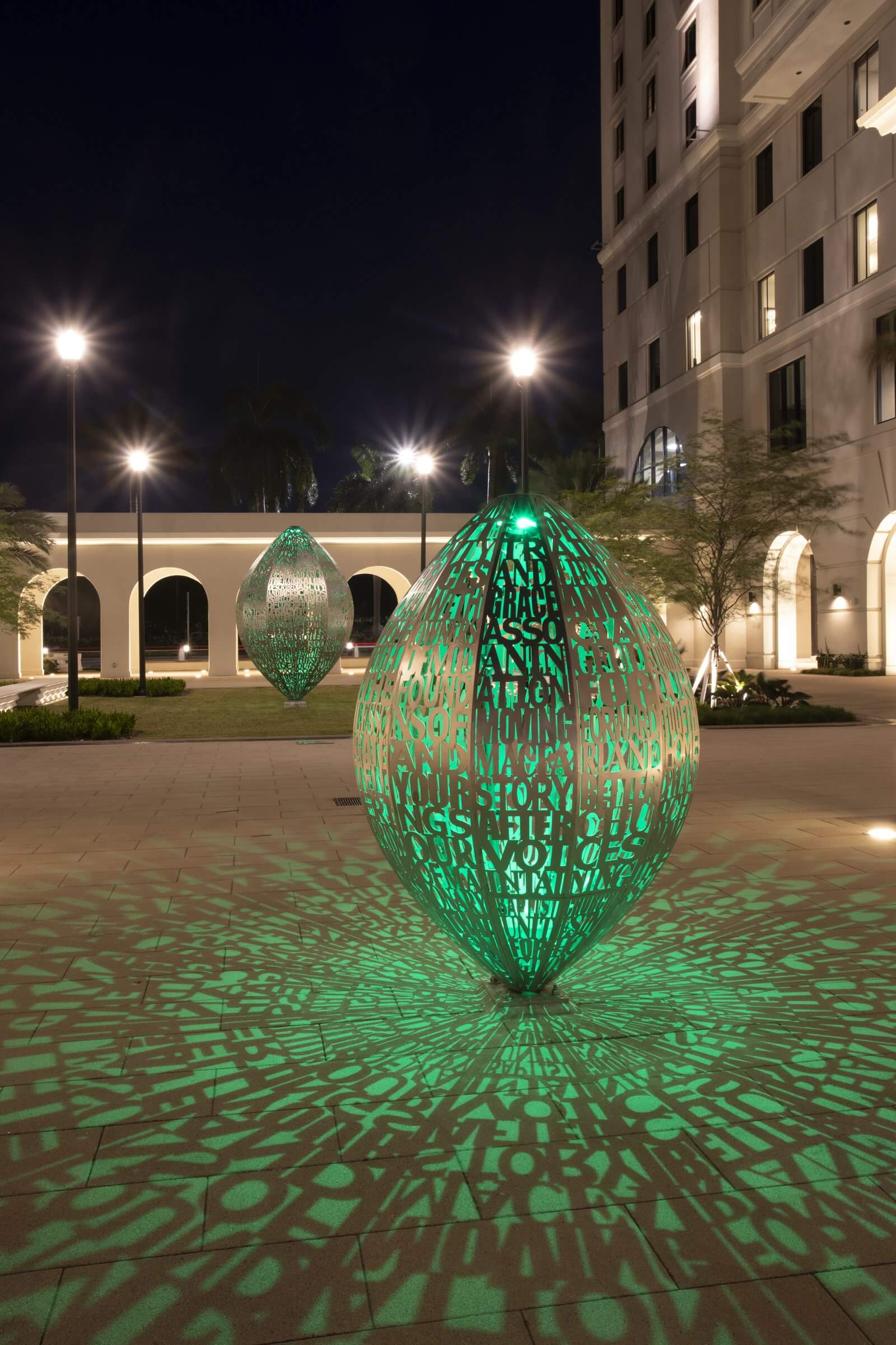 Two large, green, oval sculptures with intricate cut-out patterns by Blessing Hancock are illuminated at night in a modern courtyard. The light from the sculptures casts complex shadows on the ground. The background includes a tall building and arched pathways lit by street lamps.