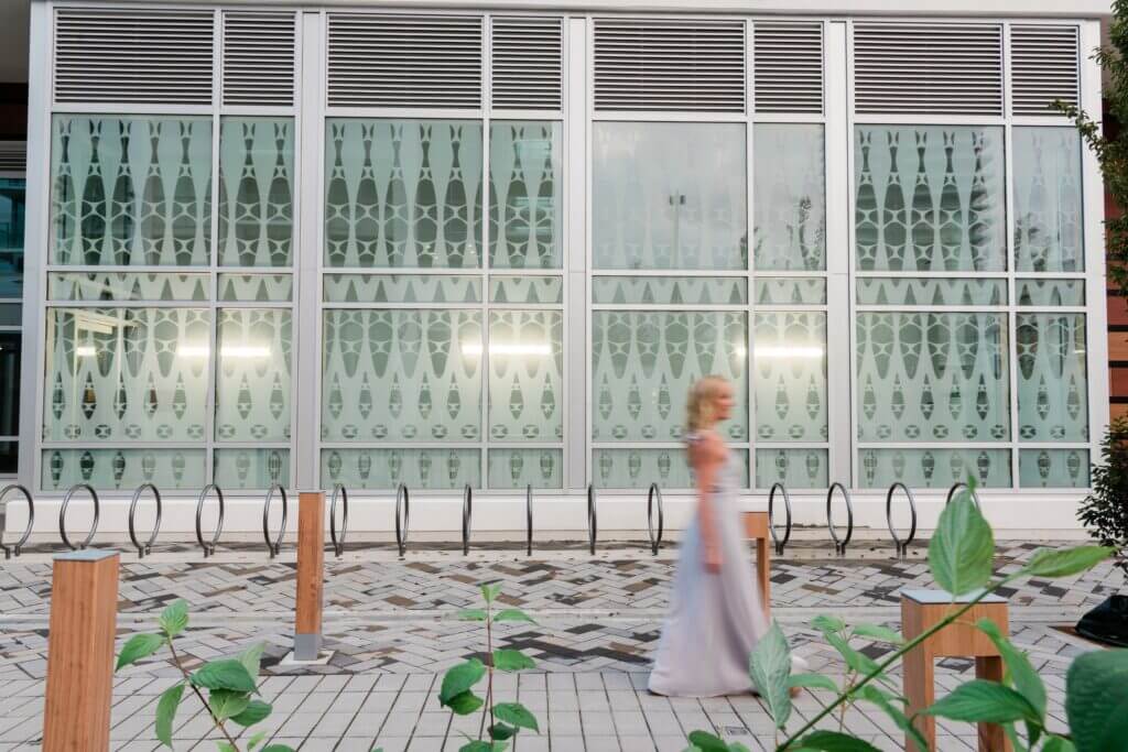 A person in a flowing white dress walks past a modern building with a large patterned glass facade. The walkway, adorned with a tiled design and bike racks, exudes urban elegance. In the foreground, green plants thrive, echoing the organic artistry of Blessing Hancock sculptures.