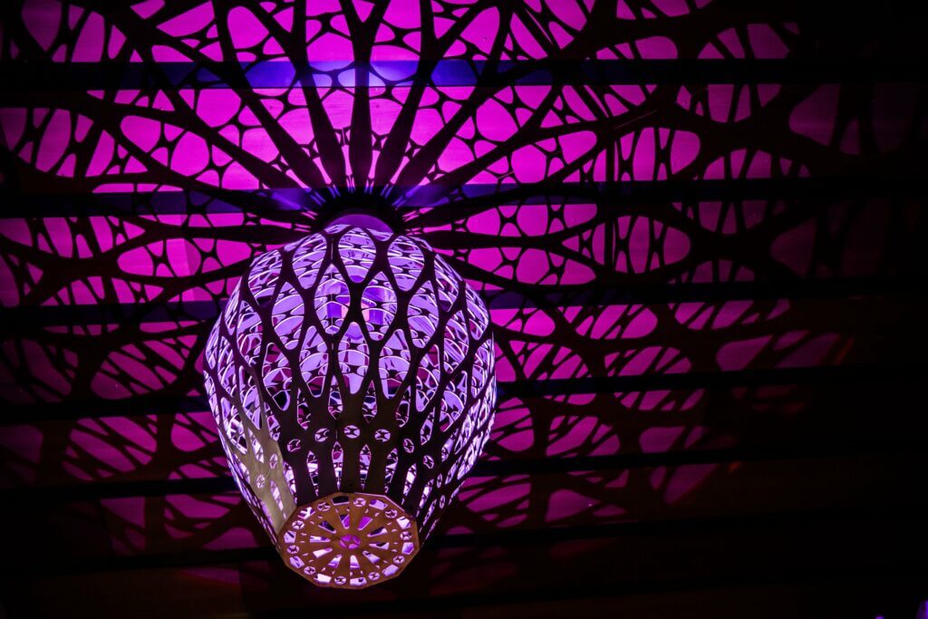 An ornate light fixture with intricate cut-out patterns casts vibrant purple and pink light onto the ceiling, creating a striking and decorative shadow effect. The design resembles a lace-like or geometric web, reminiscent of a Blessing Hancock sculpture, contributing to a modern and artistic ambiance.