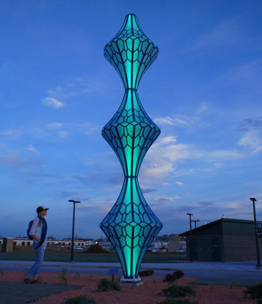 A person wearing a hat and a backpack stands next to a tall, illuminated Blessing Hancock sculpture with blue and green hues. The abstract piece has a segmented, hourglass-like shape and is set against a twilight sky with scattered clouds and a building in the background.