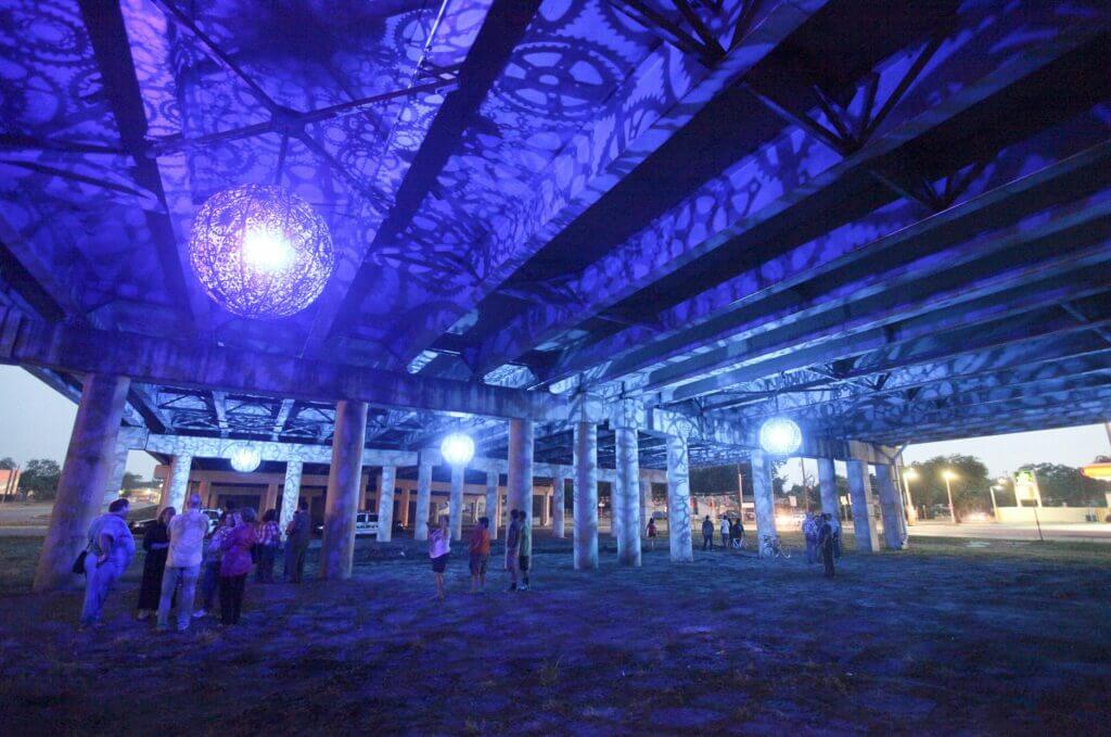 People walk under a bridge illuminated by blue and purple lights with intricate gear patterns projected onto the ceiling. The area, showcasing the creative essence of Blessing Hancock sculpture artist, is adorned with spherical light fixtures, creating an artistic and vibrant atmosphere in the evening.