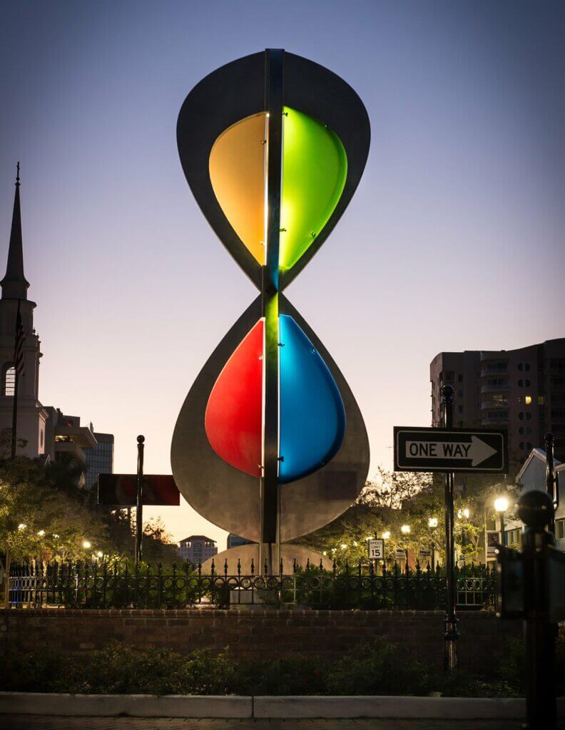 A large, colorful hourglass-shaped sculpture by artist Blessing Hancock stands illuminated at dusk against a backdrop of city buildings and a church steeple. The sculpture features vibrant sections in green, yellow, blue, and red. A "One Way" street sign is visible in the foreground.