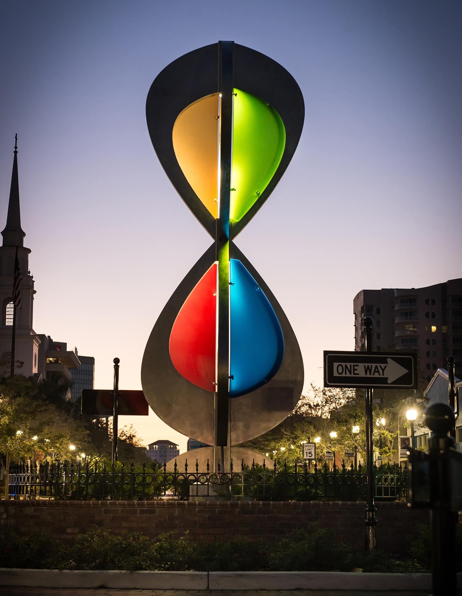 A modern sculpture by Blessing Hancock, featuring an hourglass-like form with four vibrant segments in red, green, yellow, and blue, stands illuminated at dusk. A church steeple and tall buildings fill the background, while a "One Way" street sign is visible to the right.