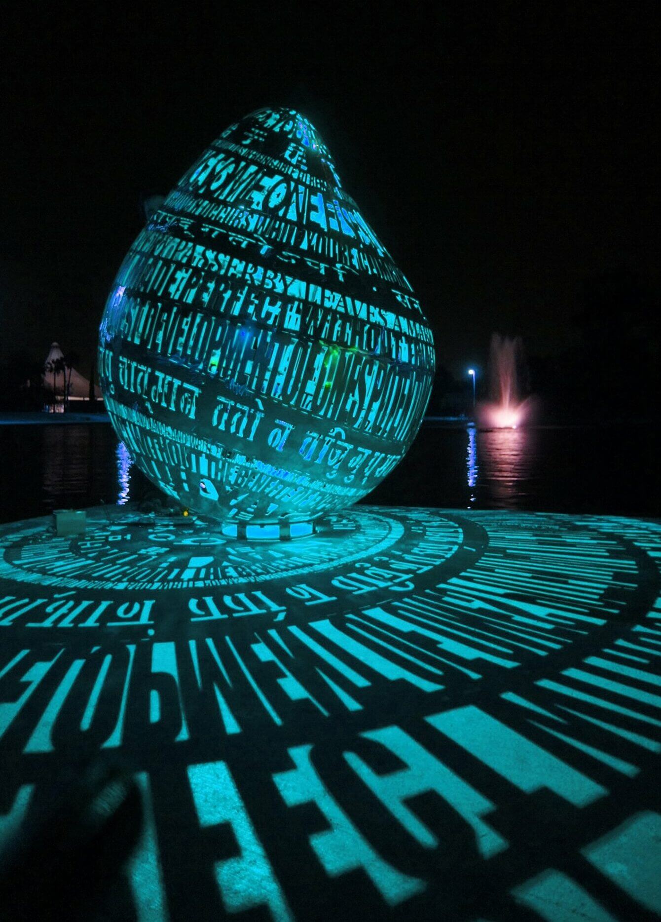 A teardrop-shaped sculpture by Blessing Hancock, illuminated in blue light, is adorned with multiple languages carved into its surface. The light casts the characters' shadows onto the ground, creating intricate patterns. A lit water fountain is visible in the background, enhancing the night scene.