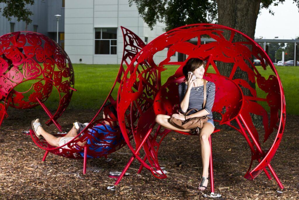 A woman sits on a vibrant red spherical metal sculpture, designed with intricate patterns by the renowned artist Blessing Hancock, in an outdoor space. Another person is lying back inside a similar sculpture nearby. Both sculptures contrast against the green grass and the modern building in the background.