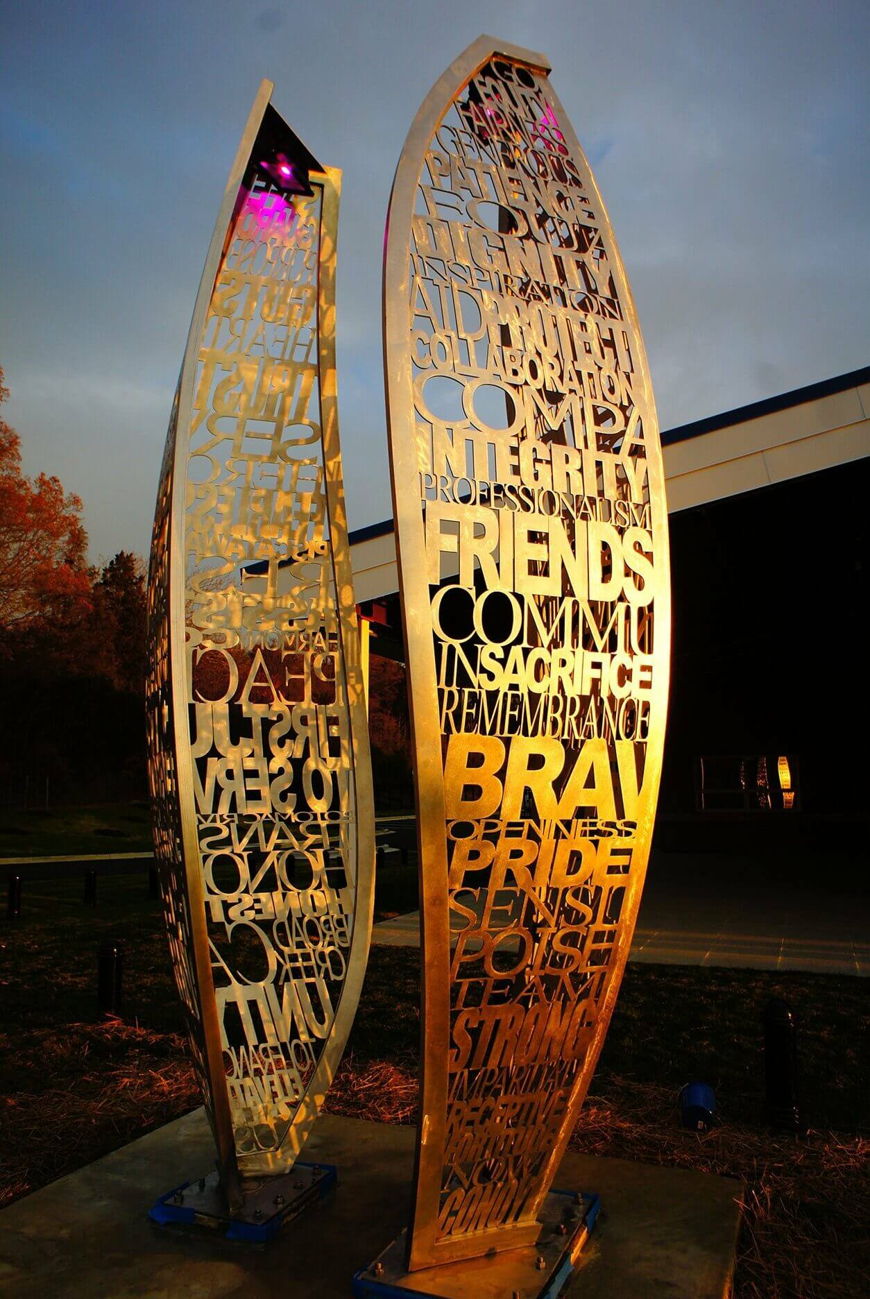 A photograph of a dual-panel metal sculpture by Blessing Hancock, with laser-cut words illuminated by golden light against a dusk sky. The panels are tall and curved, featuring words like "courage," "brave," "friends," and "sacrifice," standing outside a modern building.