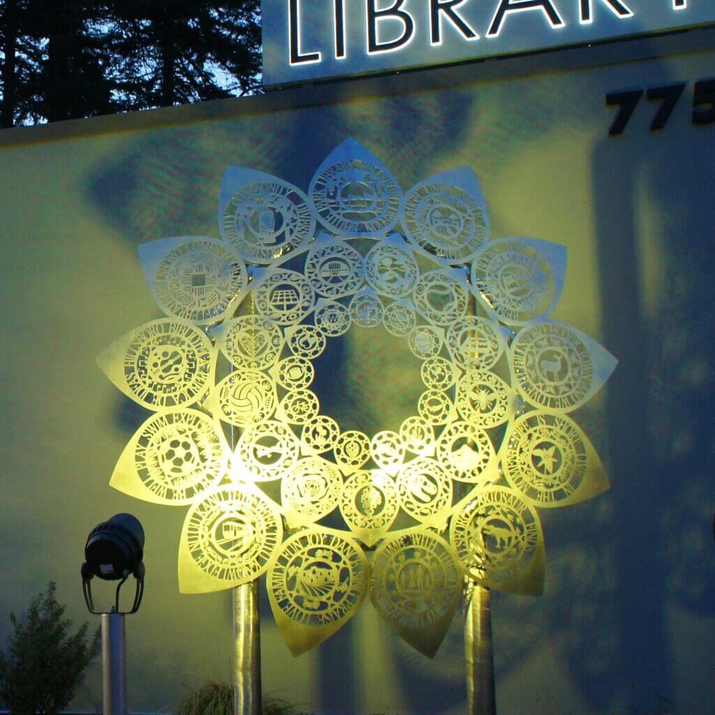 A decorative metal sculpture resembling a flower, created by the renowned artist Blessing Hancock, is illuminated at night in front of a building with the word "LIBRARY" partially visible at the top. The intricate designs are lit by a spotlight, and some numbers "775" are seen on the building's side.