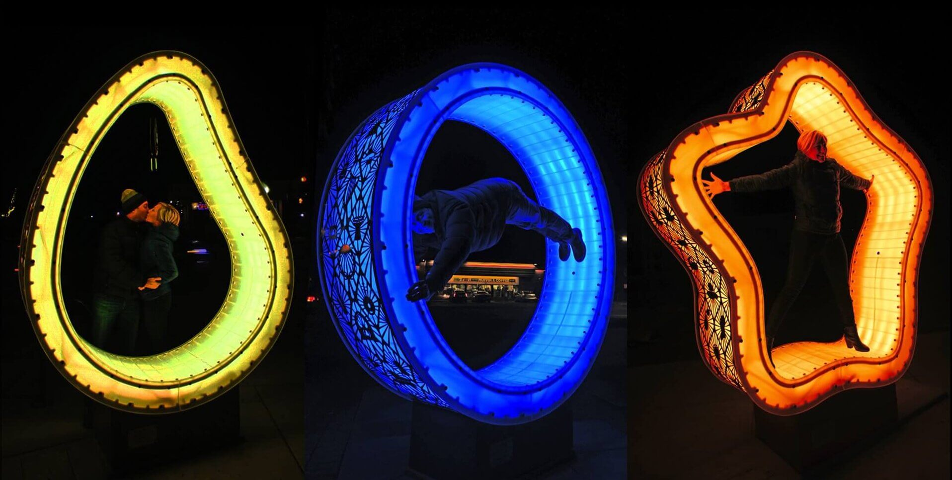 A night scene displays three brightly lit, differently shaped sculptures by artist Blessing Hancock. Each sculpture features a person posing inside. Left: a green shape resembling an avocado. Center: a blue circular shape. Right: an orange star-like shape.