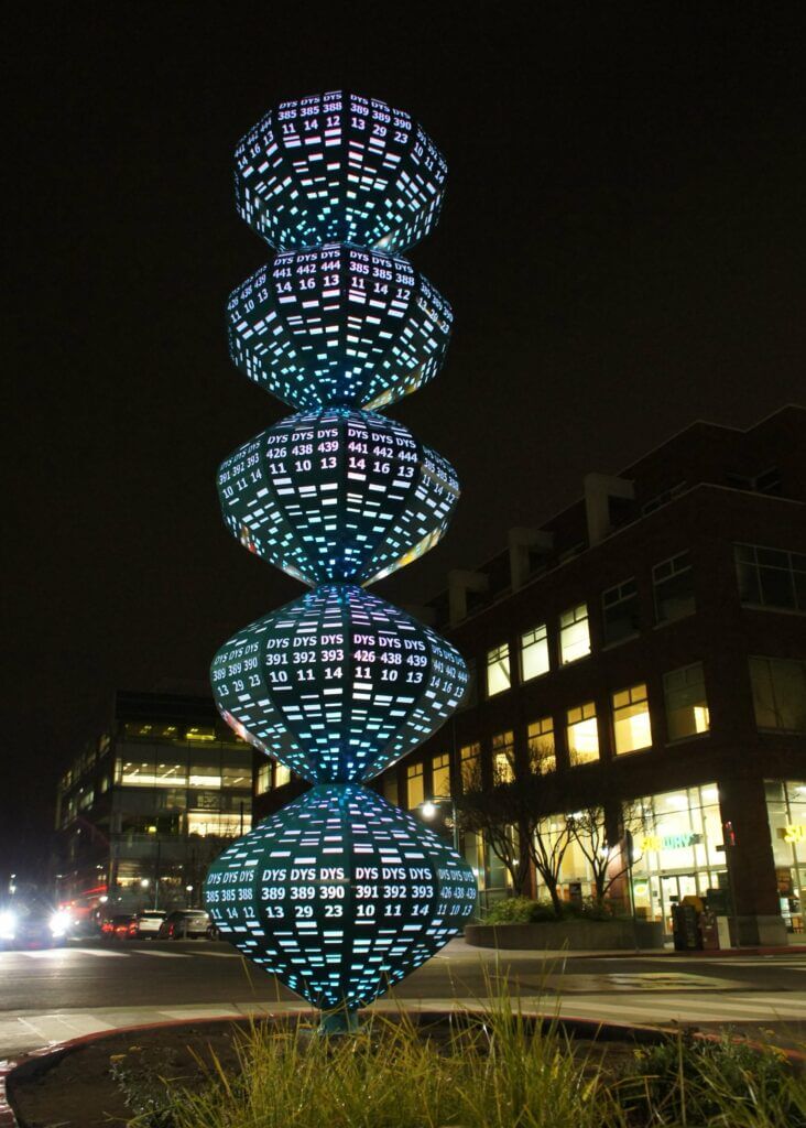 A tall, illuminated sculpture by Blessing Hancock features stacked, spherical shapes with numbers and letters glowing in various colors. The artwork is located outdoors, near modern buildings with lit windows at night.