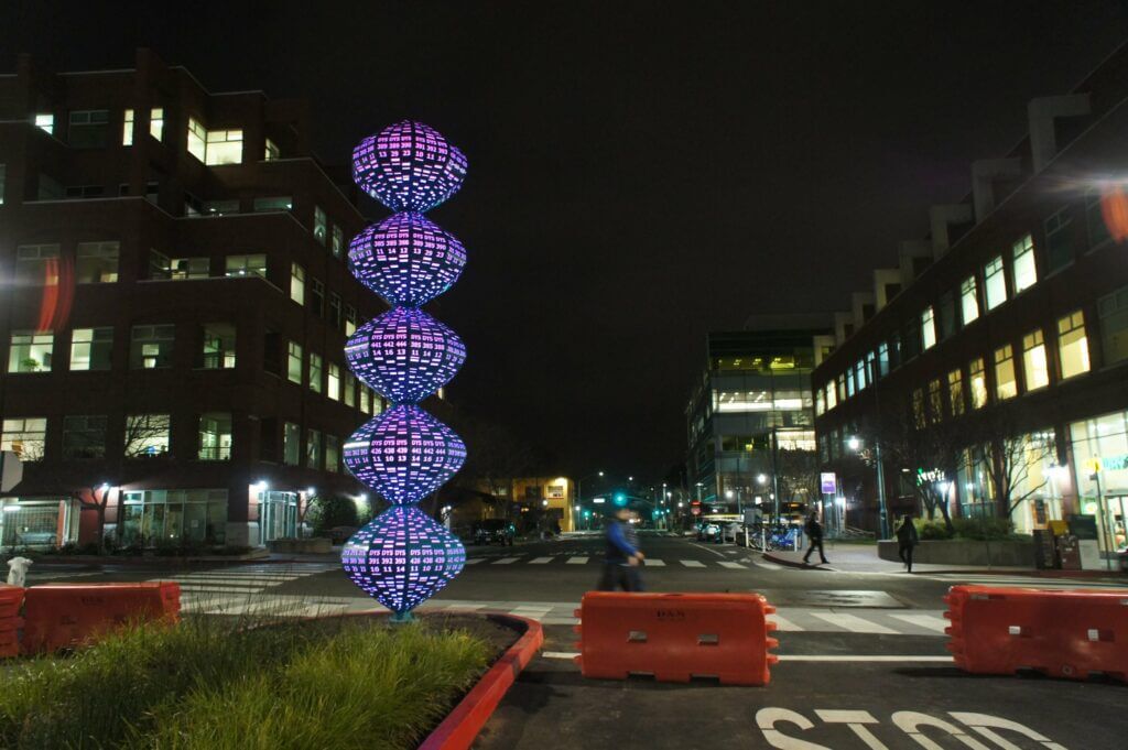 A nighttime street scene features a vertical sculpture composed of five illuminated, concentric shapes that glow purple. Created by the renowned Blessing Hancock, the sculpture is placed in the middle of an intersection lined with orange barriers. Office buildings with lit windows are in the background.