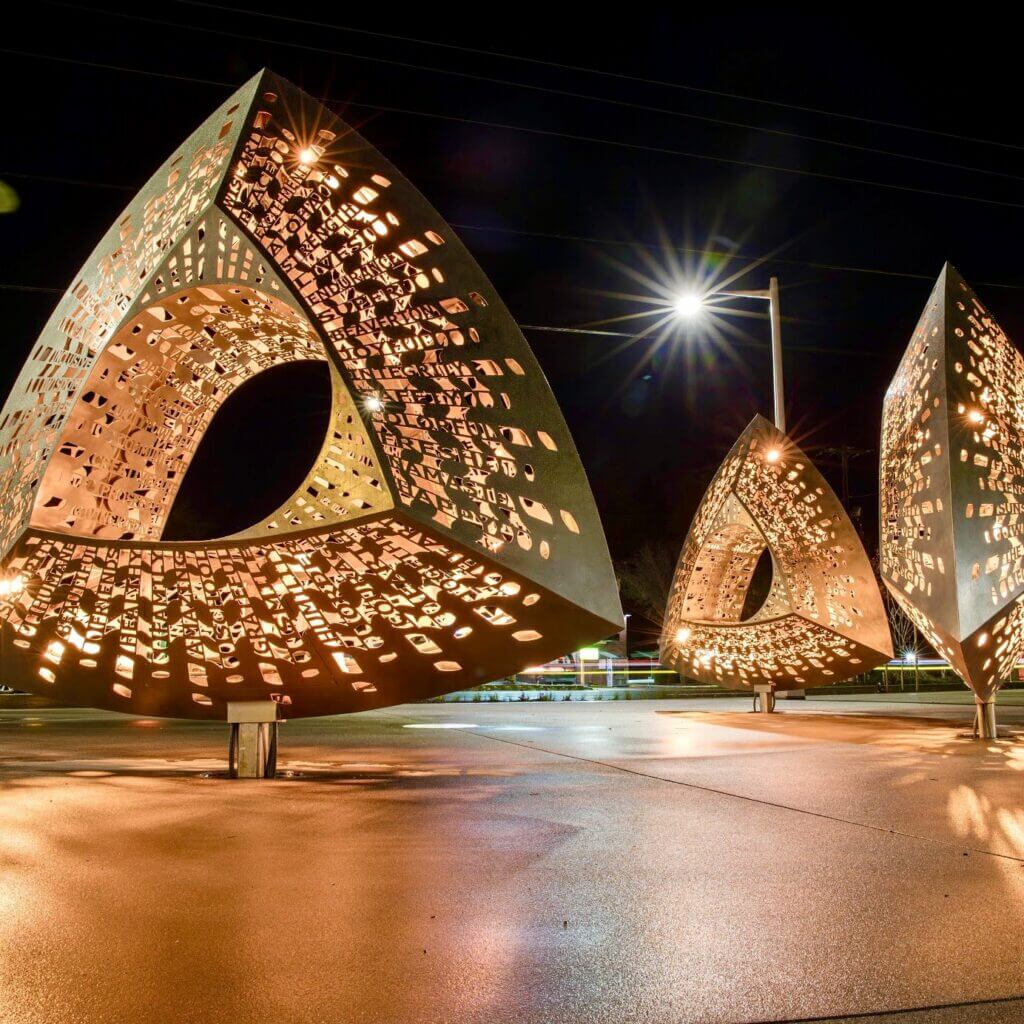 A nighttime image of two large, triangular metal sculptures with intricate cut-out designs. Created by renowned sculpture artist Blessing Hancock, the pieces are illuminated from within, casting complex shadows on the surrounding area. A streetlight shines in the background, enhancing the ambient lighting.