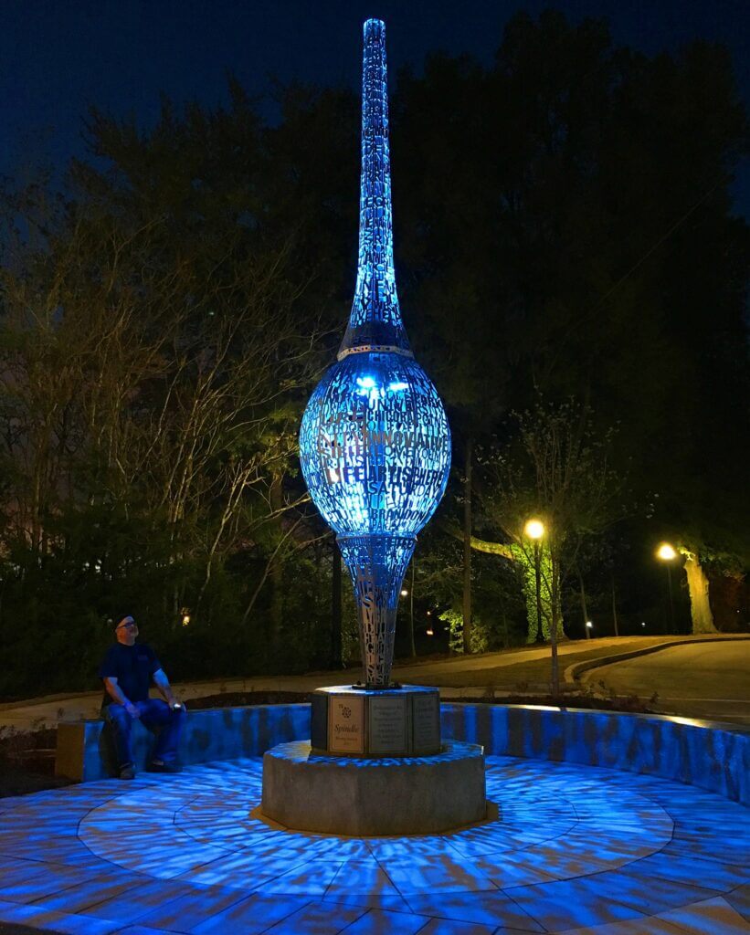 A person sits on the edge of a circular structure featuring a tall, intricately designed sculpture by acclaimed artist Blessing Hancock. Illuminated at night, it emits a blue glow, casting patterns onto the ground. Trees and streetlights can be seen in the background.