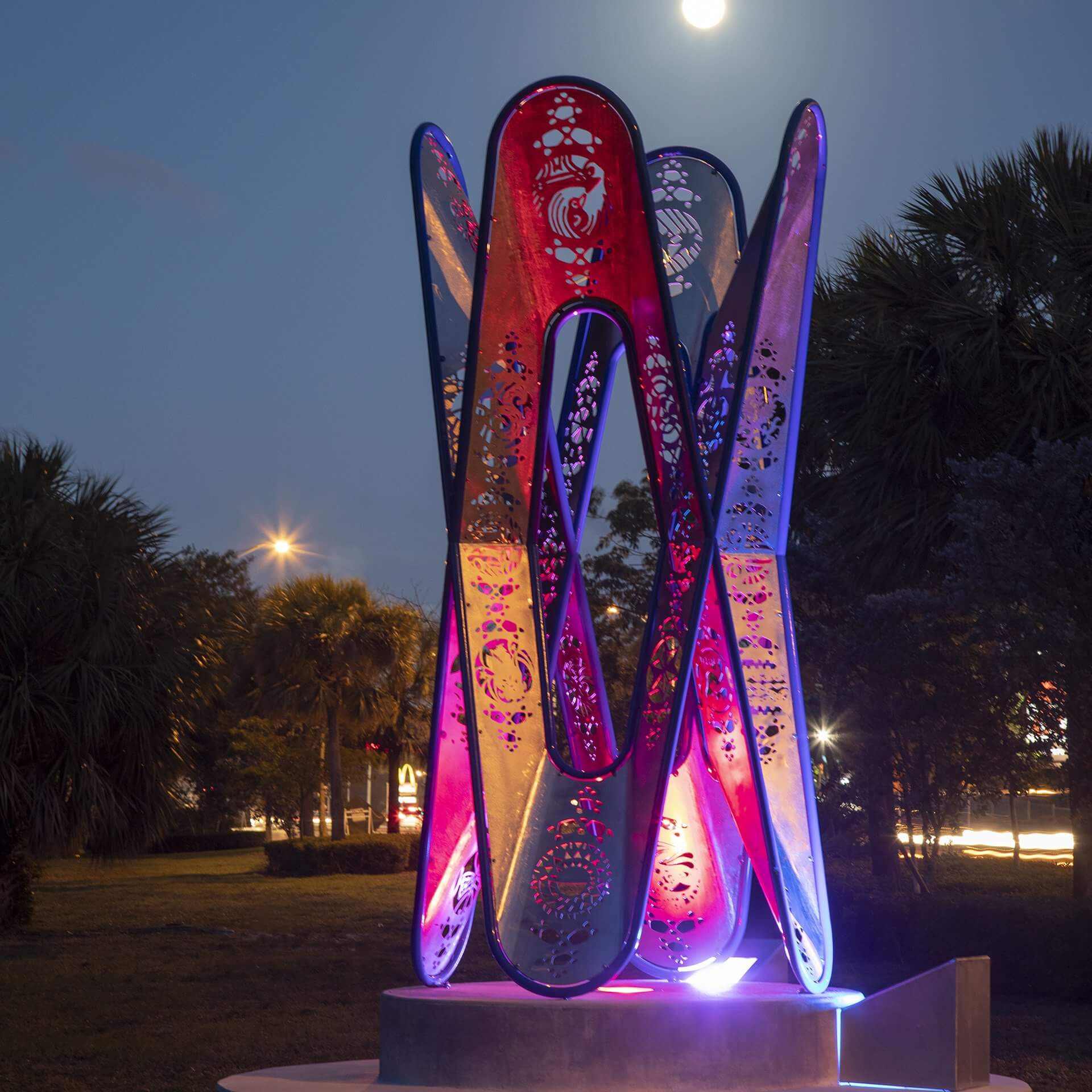 A vibrant, multicolored abstract sculpture by the renowned Blessing Hancock consists of intertwined vertical elements, illuminated against a twilight sky with a visible moon in the background. The structure stands amidst palm trees and nearby streetlights.