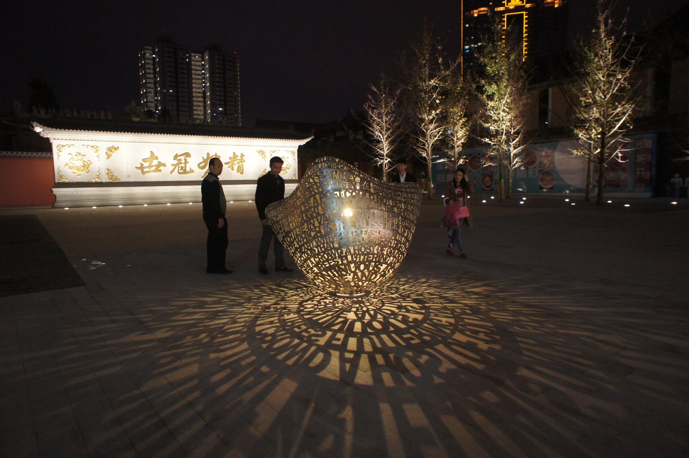 A nighttime scene features an intricate metal sculpture by Blessing Hancock, casting elaborate shadows on the pavement, illuminated by lights. In the background, several people are walking near buildings adorned with lights and colorful signs. Tall, lit buildings are partially visible.