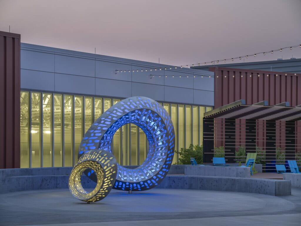 A modern art installation by renowned sculpture artist Blessing Hancock features two interlocking ring structures illuminated in blue and yellow lights. The background showcases a contemporary building with large windows and hanging string lights above, alongside blue chairs and greenery on a patio.