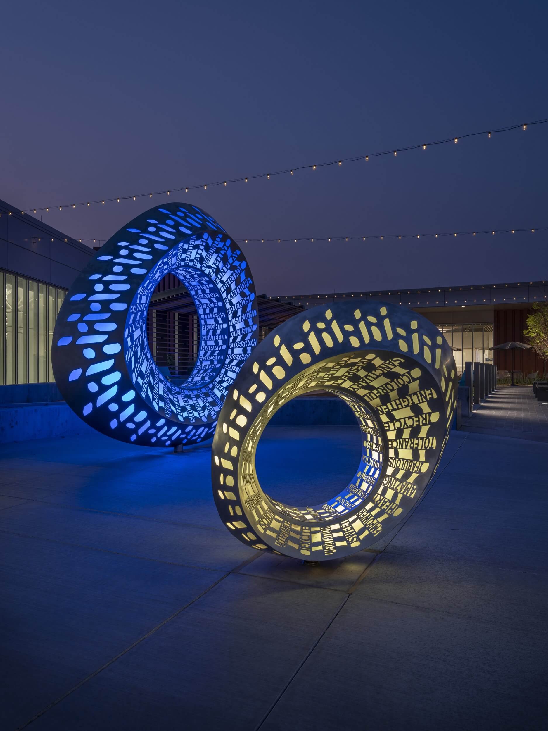 Two large, circular sculptures with intricate cutout patterns are illuminated in blue and yellow lights. Created by the renowned Blessing Hancock, the sculptures stand outdoors on a paved area, with string lights hanging overhead against the evening sky.