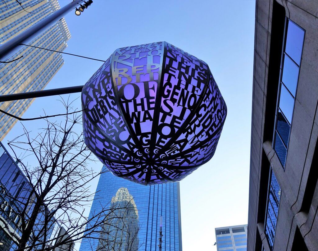 A Blessing Hancock sculpture made of metal letters forming geometric shapes is suspended high between buildings in an urban area. The sky is clear, and the reflective glass windows of the surrounding skyscrapers shine brilliantly in the background.