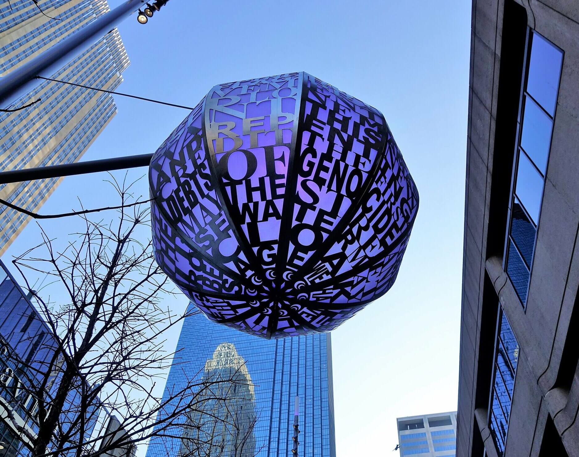 A large, geometric sculpture by Blessing Hancock is suspended in an urban environment, consisting of dark, overlapping letters forming various words. Skyscrapers reflect the blue sky in the background, while tree branches frame the sculpture from beneath.