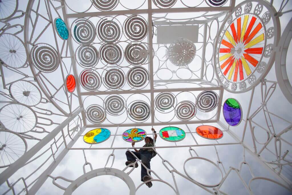 A person climbs inside an intricate metal structure filled with colorful circular decorations, spiral patterns, and bicycle wheels. The sky is overcast, providing a soft, diffused light. The vibrant colors and unique design of this Blessing Hancock sculpture create an artistic atmosphere.