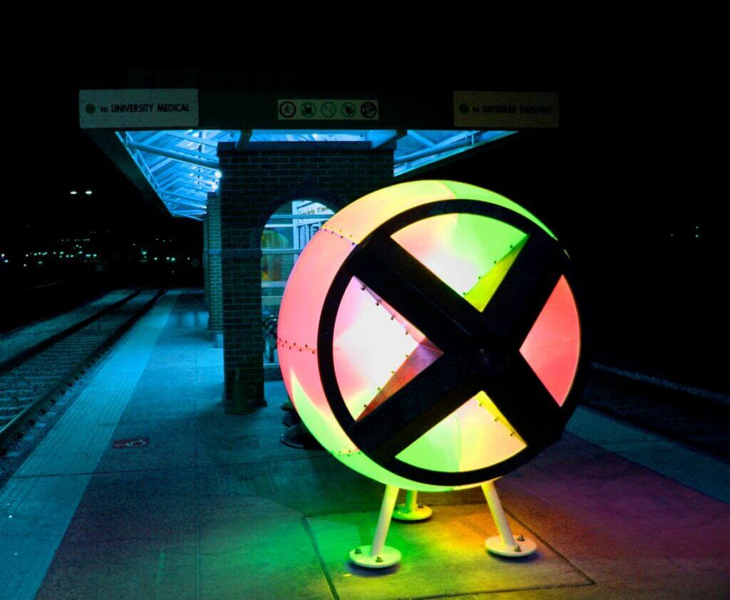 A brightly illuminated sculpture by artist Blessing Hancock, featuring multicolored lights, sits on a train station platform at night. The circular piece has an "X"-shaped design inside. The station includes a covered waiting area with a roof and seats, while signs above indicate directions.