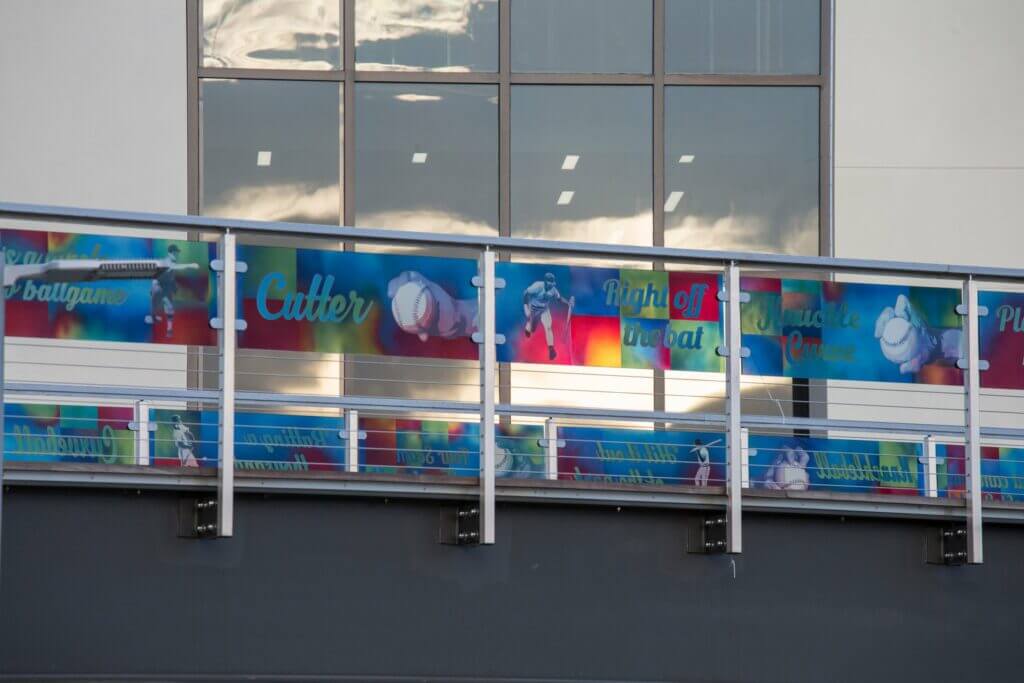 A bright, colorful outdoor banner featuring baseball-themed graphics and text is displayed on a glass railing. The reflected light on the windows above adds a dynamic touch, reminiscent of a Blessing Hancock sculpture. The words "Cutter," "Right off the bat," and "muscle curve" are visible among other text and images.