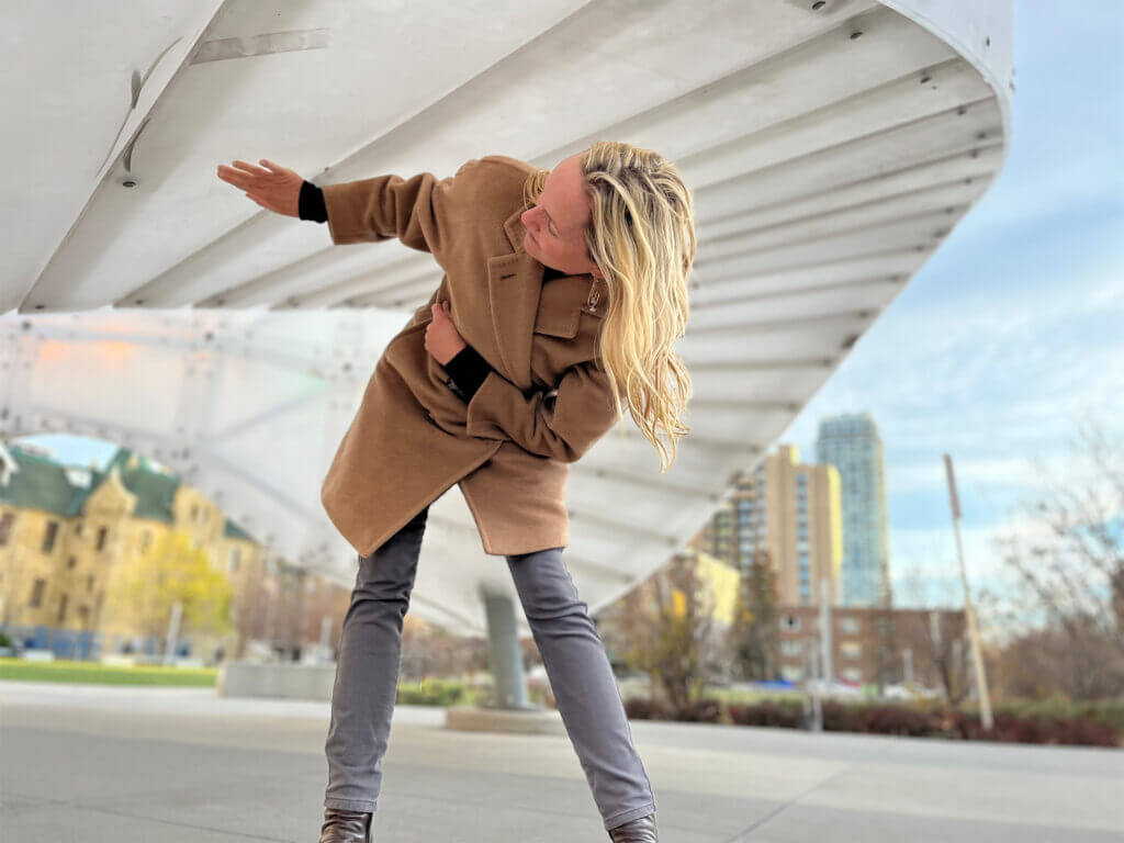 Blessing Hancock, wearing a brown coat and gray pants, leans and looks underneath a large, curved architectural structure by sculpture artist Blessing Hancock in an urban setting. Skyscrapers and trees with autumn foliage are visible in the background.