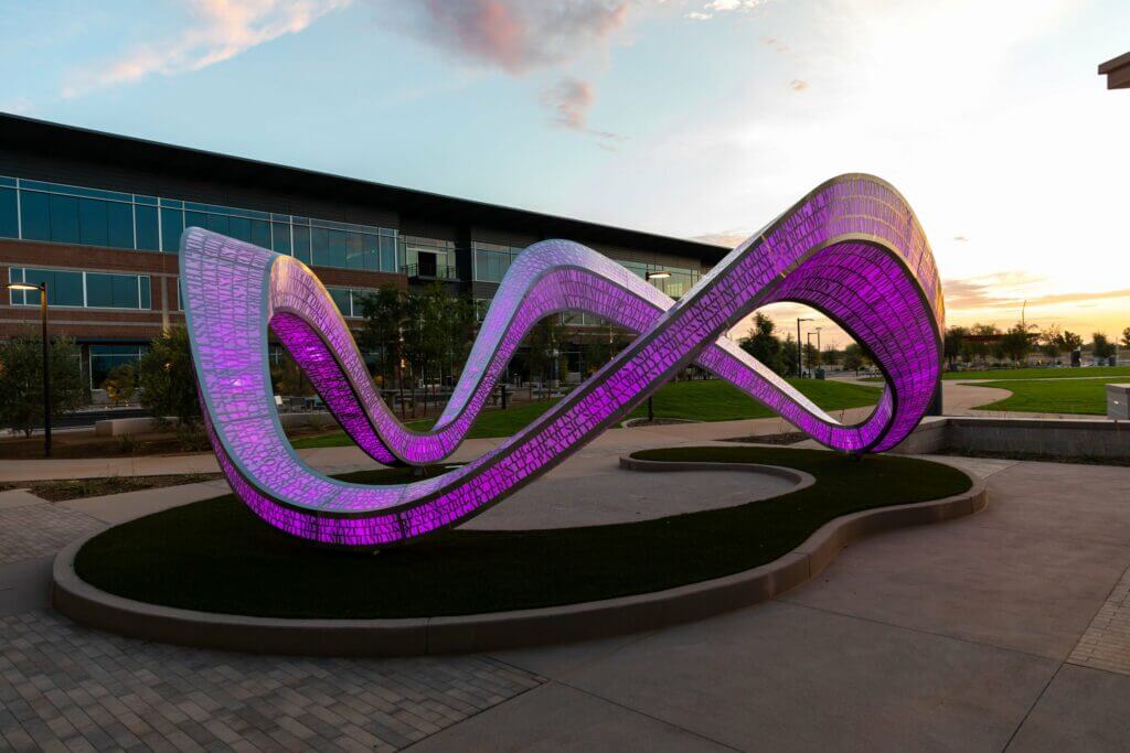 A large, illuminated Blessing Hancock sculpture featuring interwoven, wavy loops stands in an outdoor plaza. The sculpture emits a vibrant purple hue and is positioned near a modern building. The surrounding area is landscaped with grass and paved walkways, set against a sunset sky.