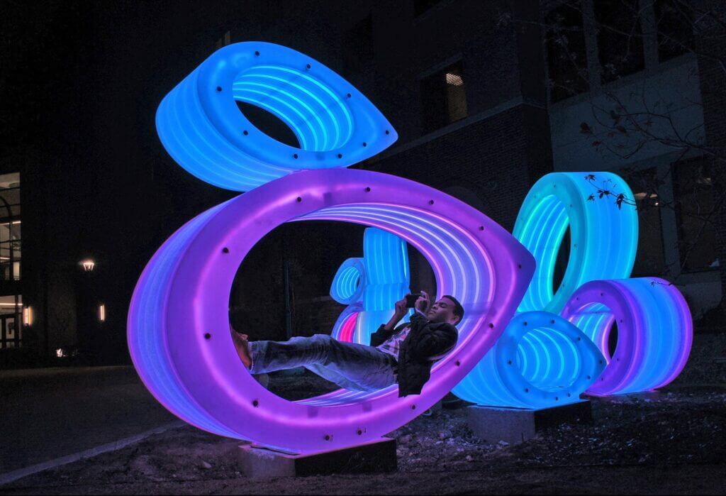 A person lounges inside a large, glowing neon sculpture at night. The Blessing Hancock sculpture is composed of interconnected circular shapes illuminated in vibrant colors including pink, blue, and purple. A building with lit windows is visible in the background.