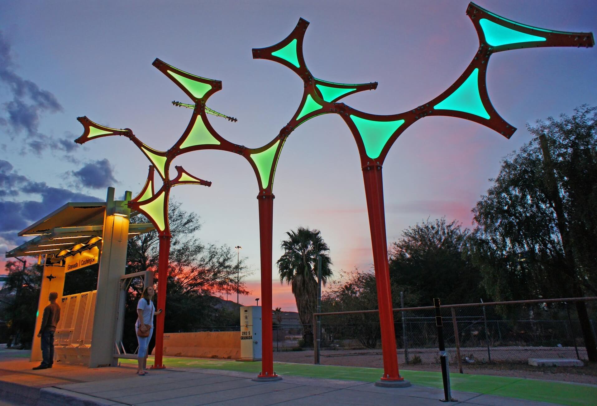 A bus stop with illuminated, artistic canopy structures in green and blue shades during sunset. Two people are waiting under the canopy, designed by renowned sculpture artist Blessing Hancock, and palm trees are visible in the background along with a colorful sky.