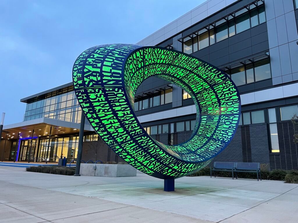 A large, modern sculpture resembling a loop by Blessing Hancock is situated in front of a contemporary building. The sculpture is adorned with green, patterned text and symbols on a dark background. The building features large windows and a sleek design. The sky is overcast.