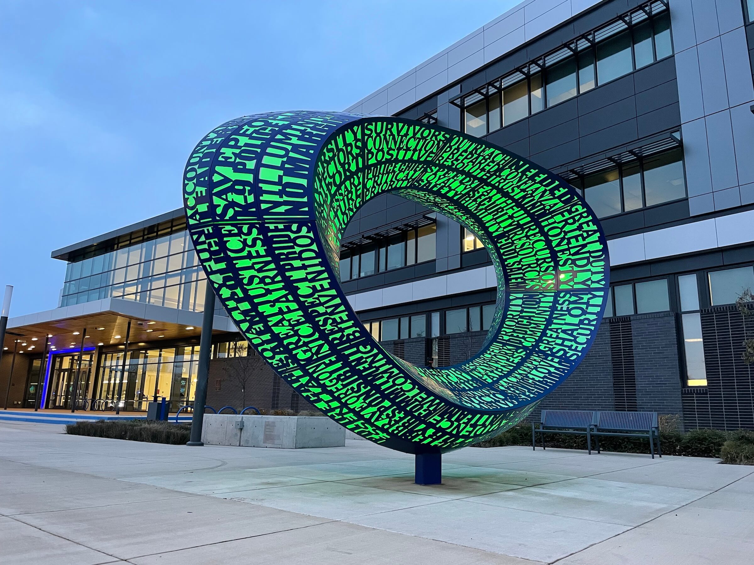 A large, modern sculpture resembling a loop by Blessing Hancock is situated in front of a contemporary building. The sculpture is adorned with green, patterned text and symbols on a dark background. The building features large windows and a sleek design. The sky is overcast.