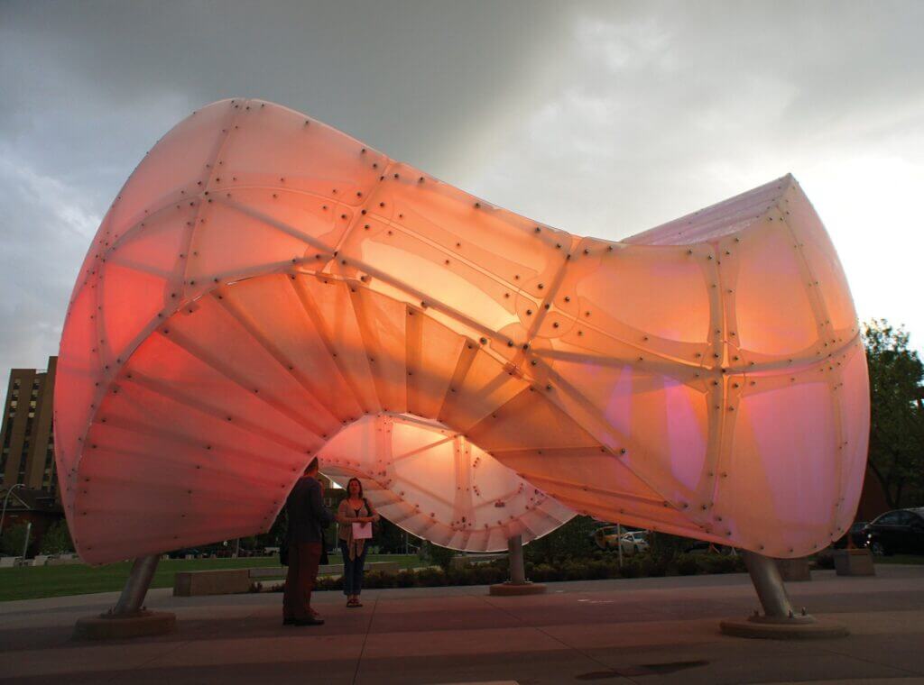 An innovative, large, translucent Blessing Hancock sculpture with an organic, twisting form is situated outdoors, illuminated from within with soft, warm lights. Two people are standing beneath the structure, conversing, with an overcast sky in the background.