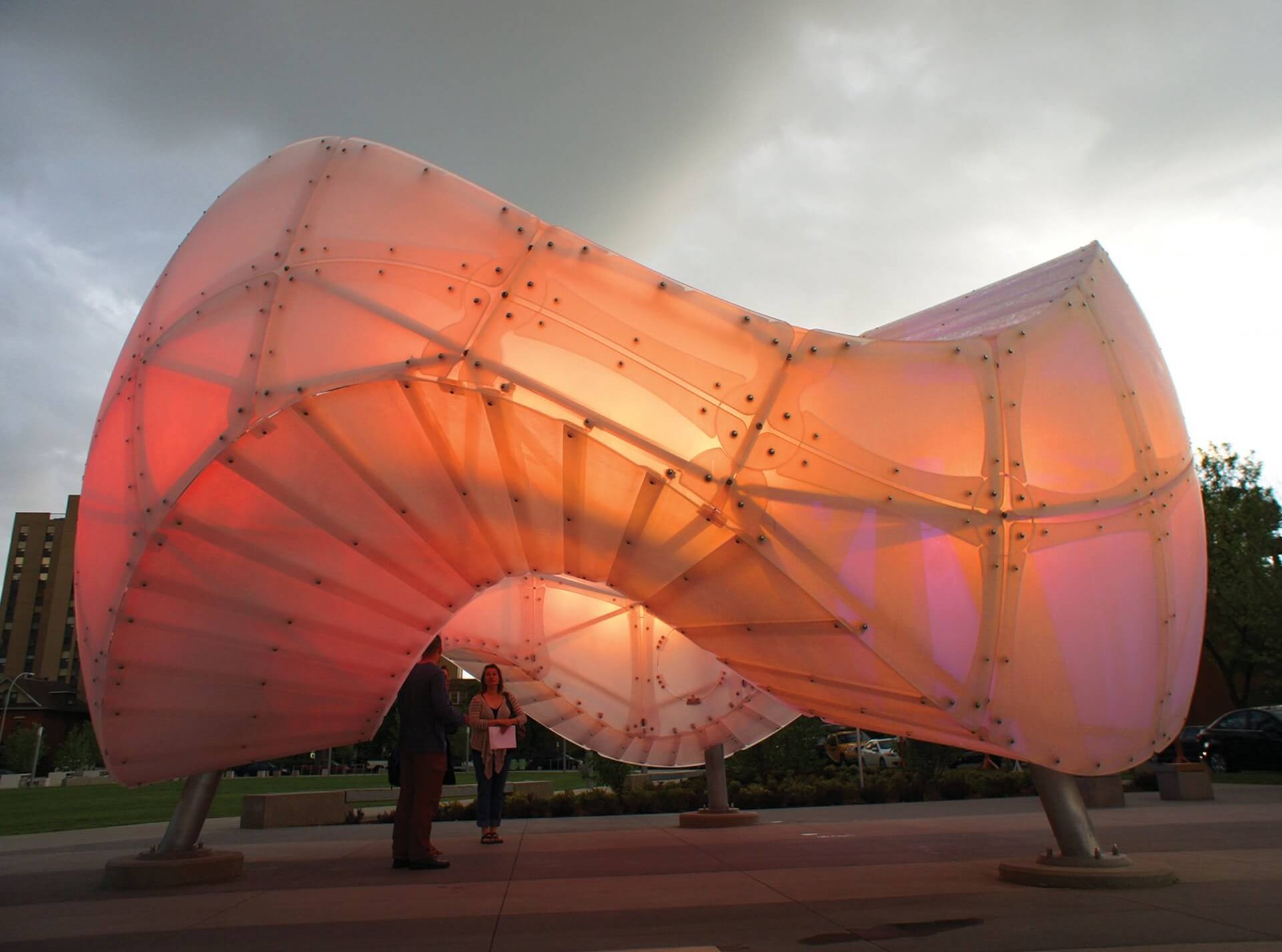 An innovative, large, translucent Blessing Hancock sculpture with an organic, twisting form is situated outdoors, illuminated from within with soft, warm lights. Two people are standing beneath the structure, conversing, with an overcast sky in the background.