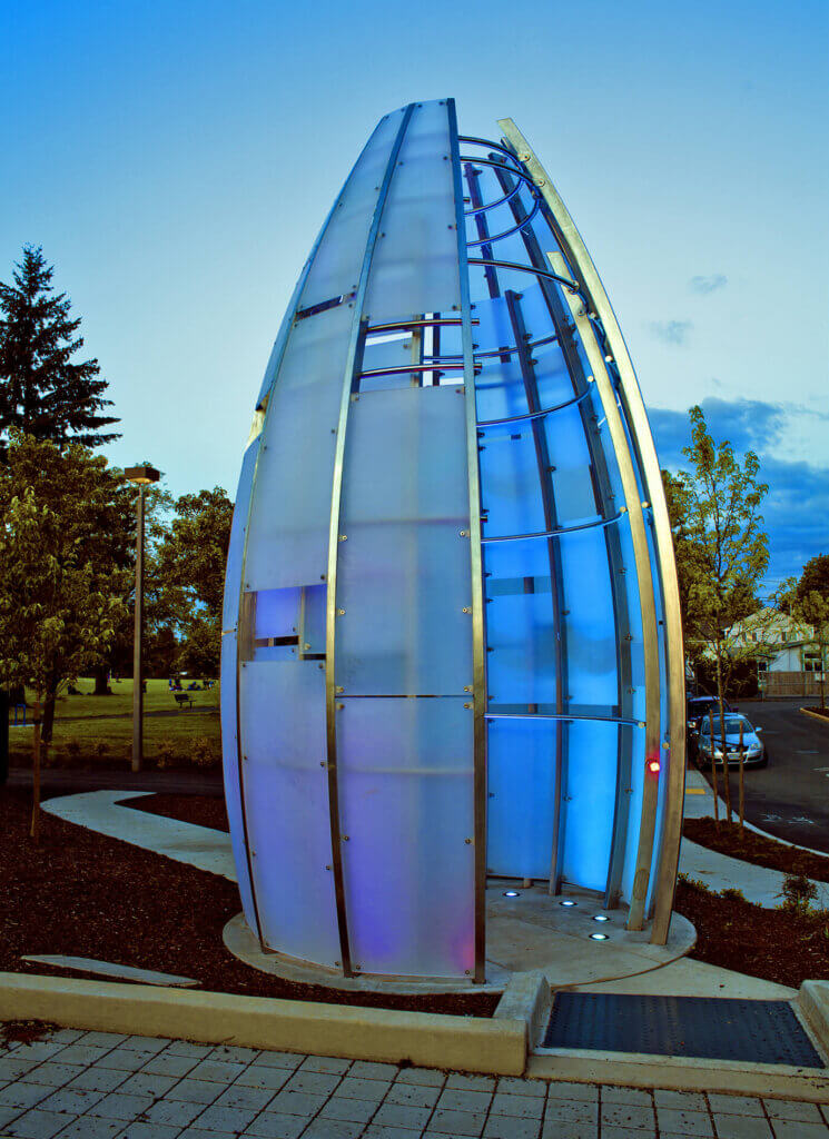 A modern, egg-shaped sculpture with translucent panels projects a mesmerizing glow in blue and purple hues against the twilight sky. Nestled on a landscaped area with trees and a pathway, it stands out while the street forms a subtle backdrop.