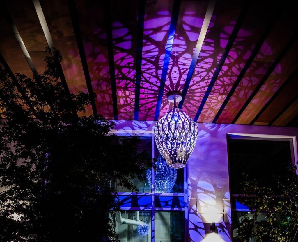 A decorative hanging light fixture projects intricate blue and purple shadows on the ceiling of a modern room. Tall windows reflect the dazzling light pattern, and part of a tree is visible on the left.