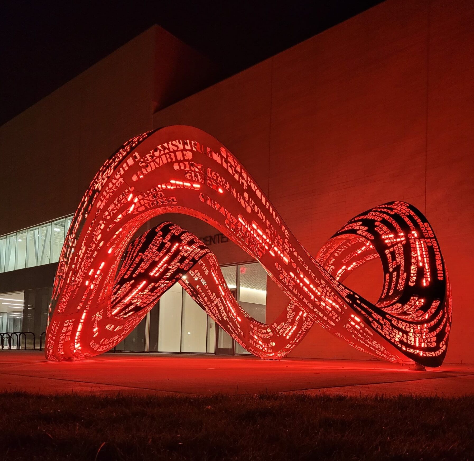 A glowing red metal sculpture with intricate cut-out text is lit at night, projecting its mesmerizing design. It forms a looping, wave-like shape, set outdoors in front of a modern building with large windows.