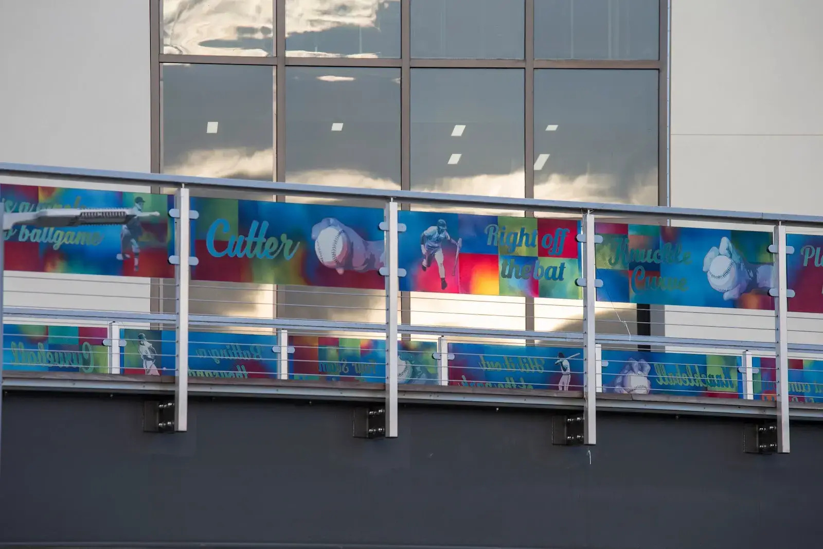 A colorful banner on a building's exterior projects baseball-themed words and images, including baseballs and players. The background features partly reflective windows showing the sky and clouds.