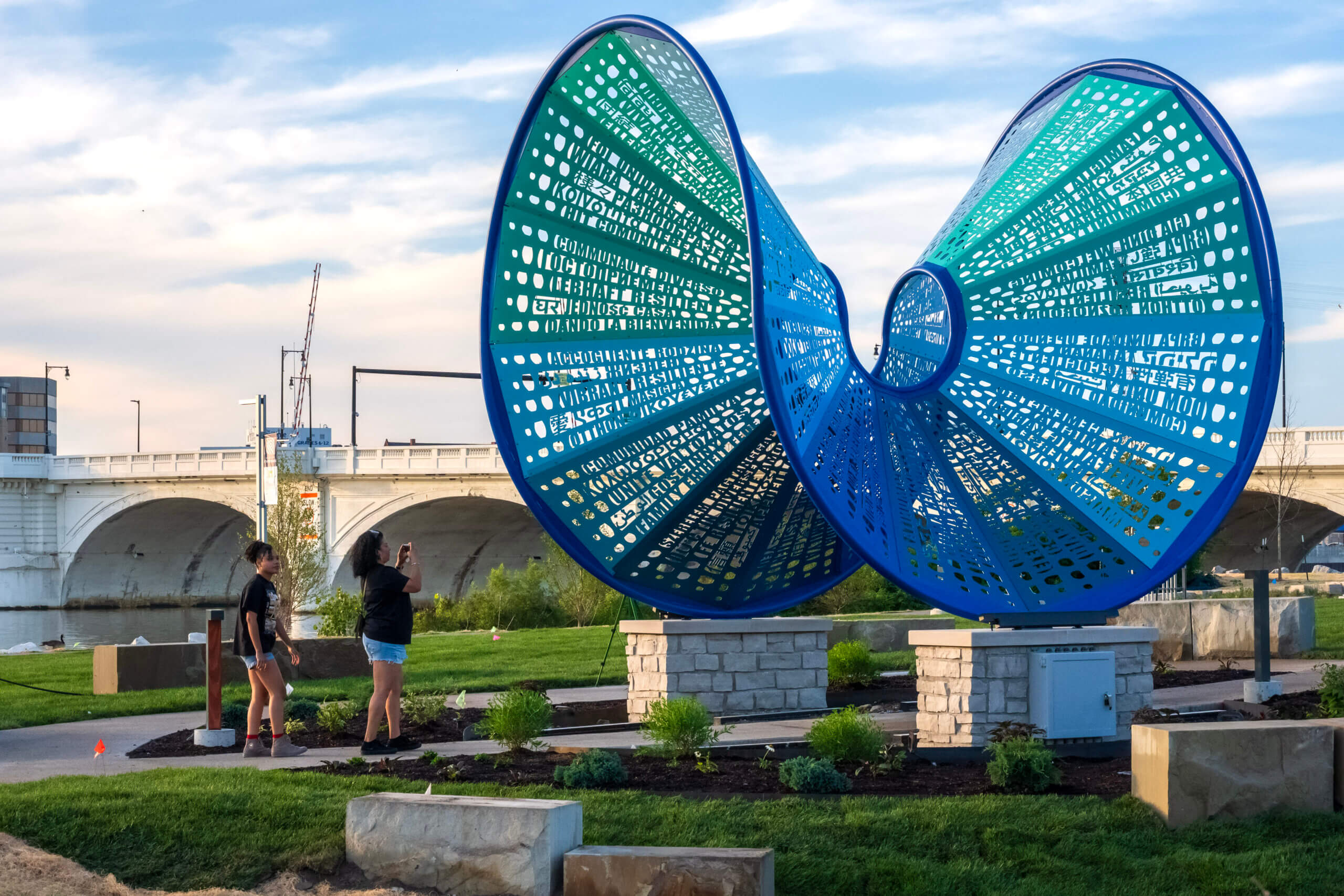 Two people stand near a large blue and green abstract metal sculpture in a park, its intricate design reminiscent of papel picado. In the background, a bridge and construction crane rise beneath the partly cloudy sky.
