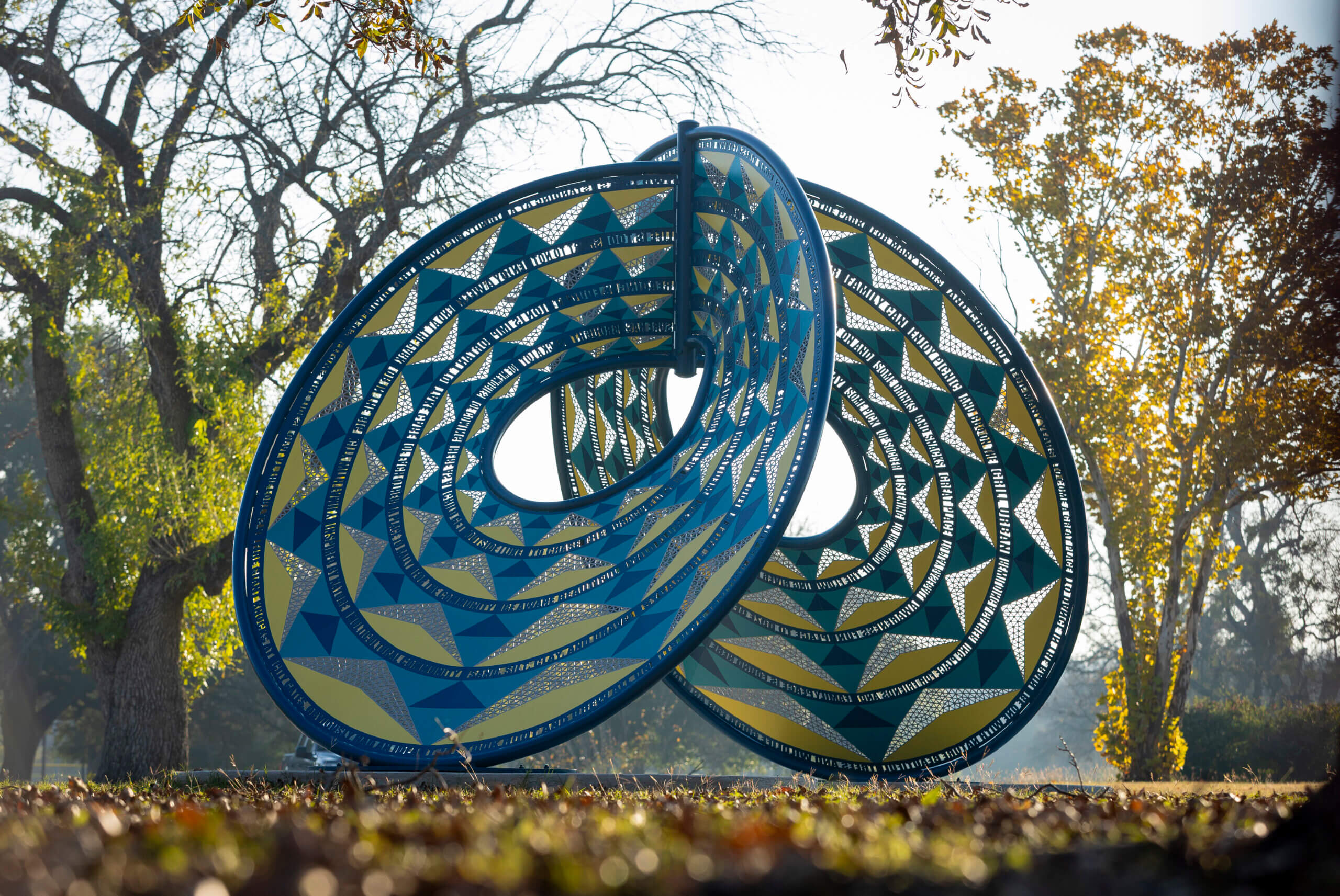 A large outdoor sculpture, crafted with Elementor precision, features two interlocking blue and yellow geometric rings with triangular patterns, set in a park where sunlight filters through leafy trees.